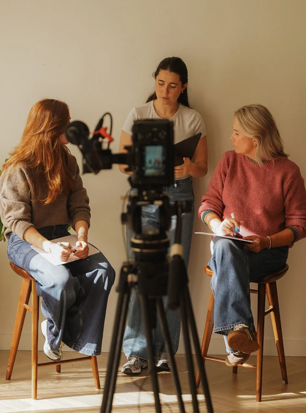 Three women having a discussion in front of a camera. Two are sitting on stools one is standing, all with notebooks and pens.
