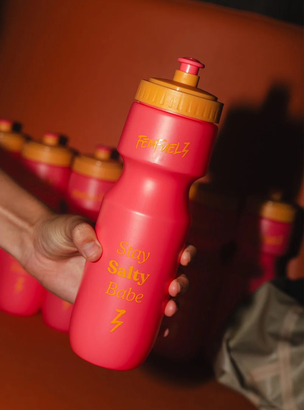 Pink water bottle with yellow lid held in hand, with repeat bottles in the background. The bottle has the text "FEMFUELZ" at the top and "Stay Salty Babe" along the middle, along with a lightning bolt symbol.