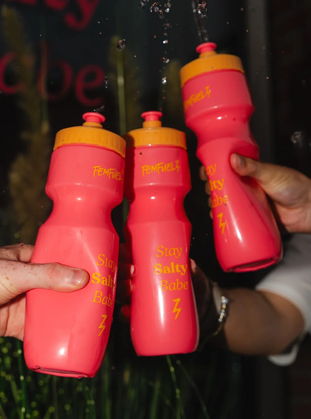 Three pink water bottles with yellow caps, held by two hands, with the phrase 'Stay Salty Babe' written in yellow on each bottle.