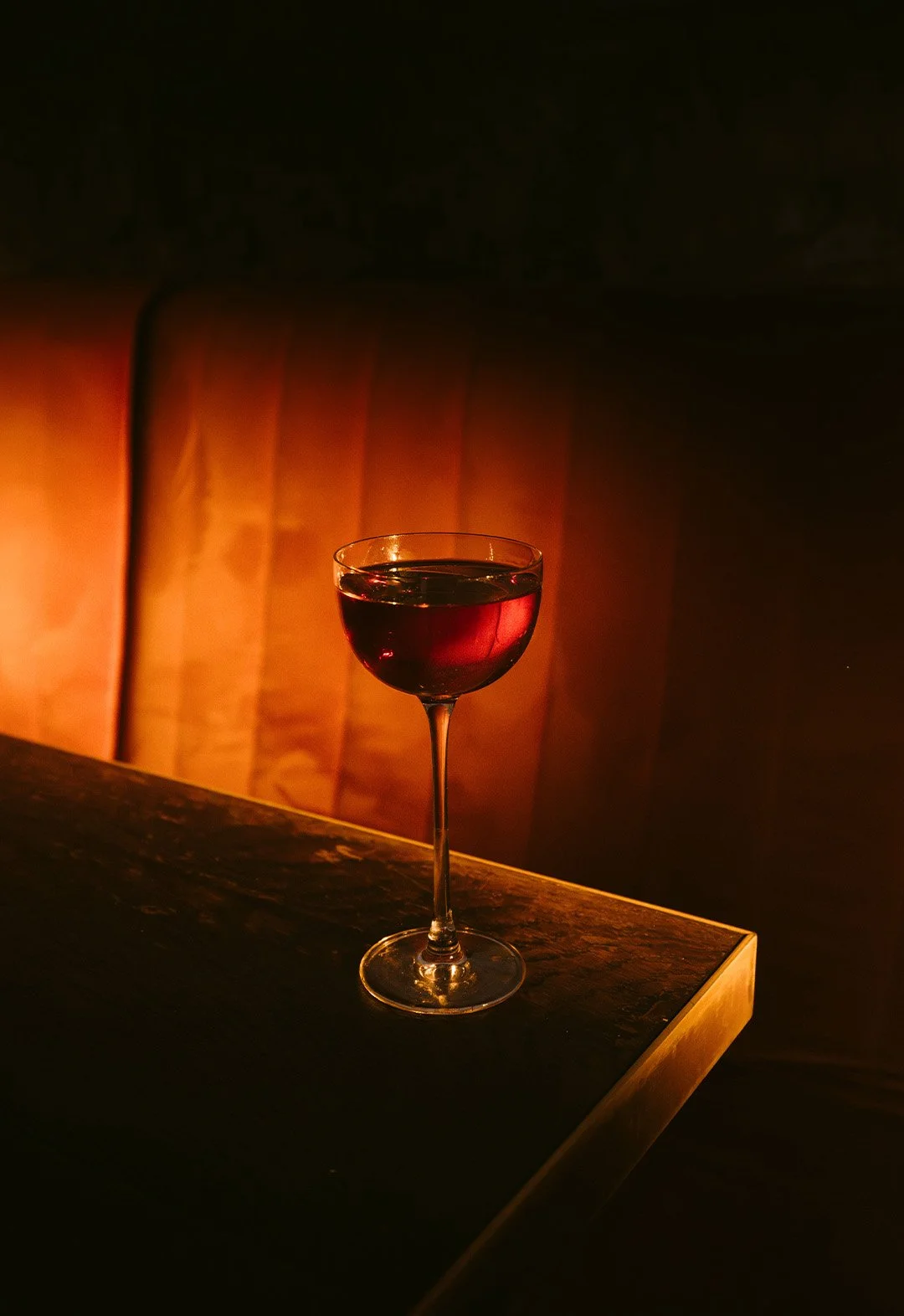 A glass of red wine on a dark wooden table, with a warm, softly lit upholstered background.