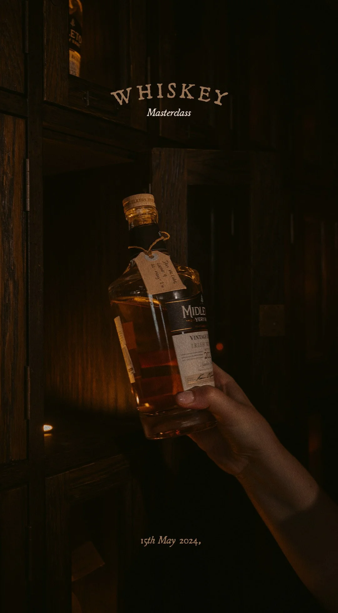A hand holding a bottle of whiskey in front of wooden shelves with alcohol bottles, with the overlay text 'WHISKEY Masterclass' and the date '15th May 2024'.