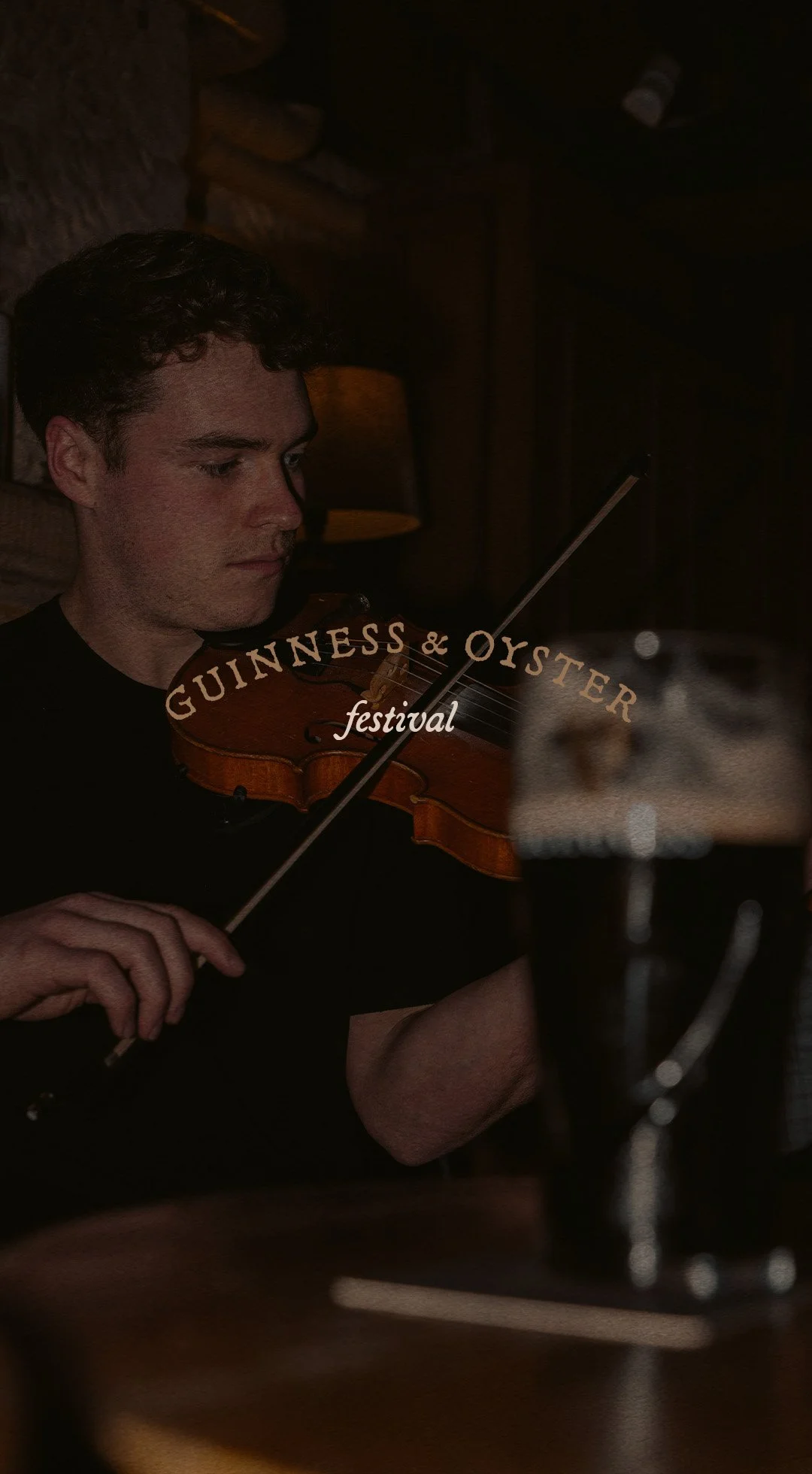 A young man with dark hair playing a violin in a dimly lit pub or music venue. There is a pint of dark beer in the foreground and the logo of the Guinness & Oyster Festival overlayed on the image.
