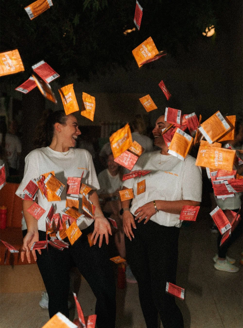 Two women laughing and having fun at a party with orange and red snack pouches falling around them.