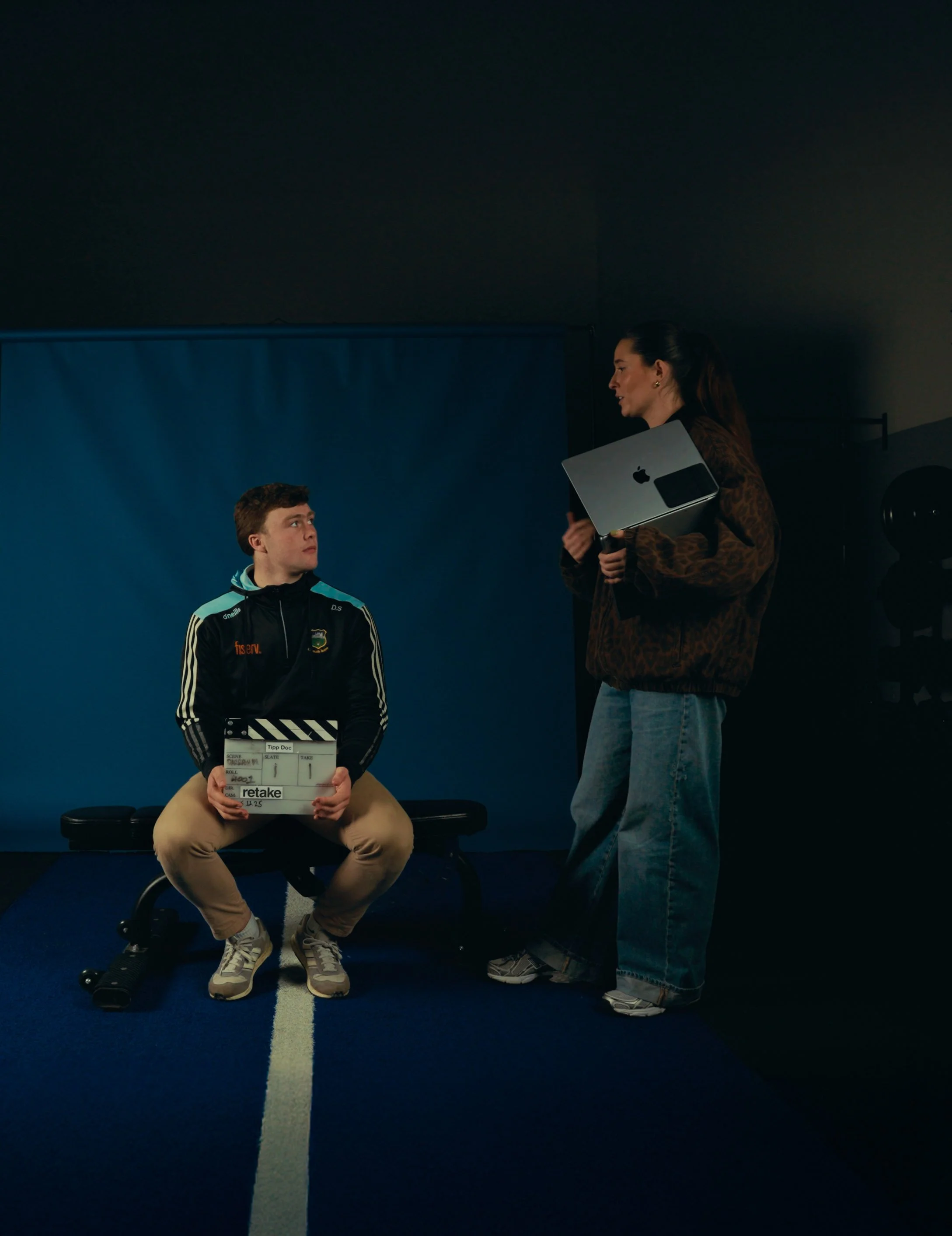 A woman with a MacBook and holding a clipboard is talking to a young man sitting on a bench, holding a clapperboard, on a blue floor with a dark background.