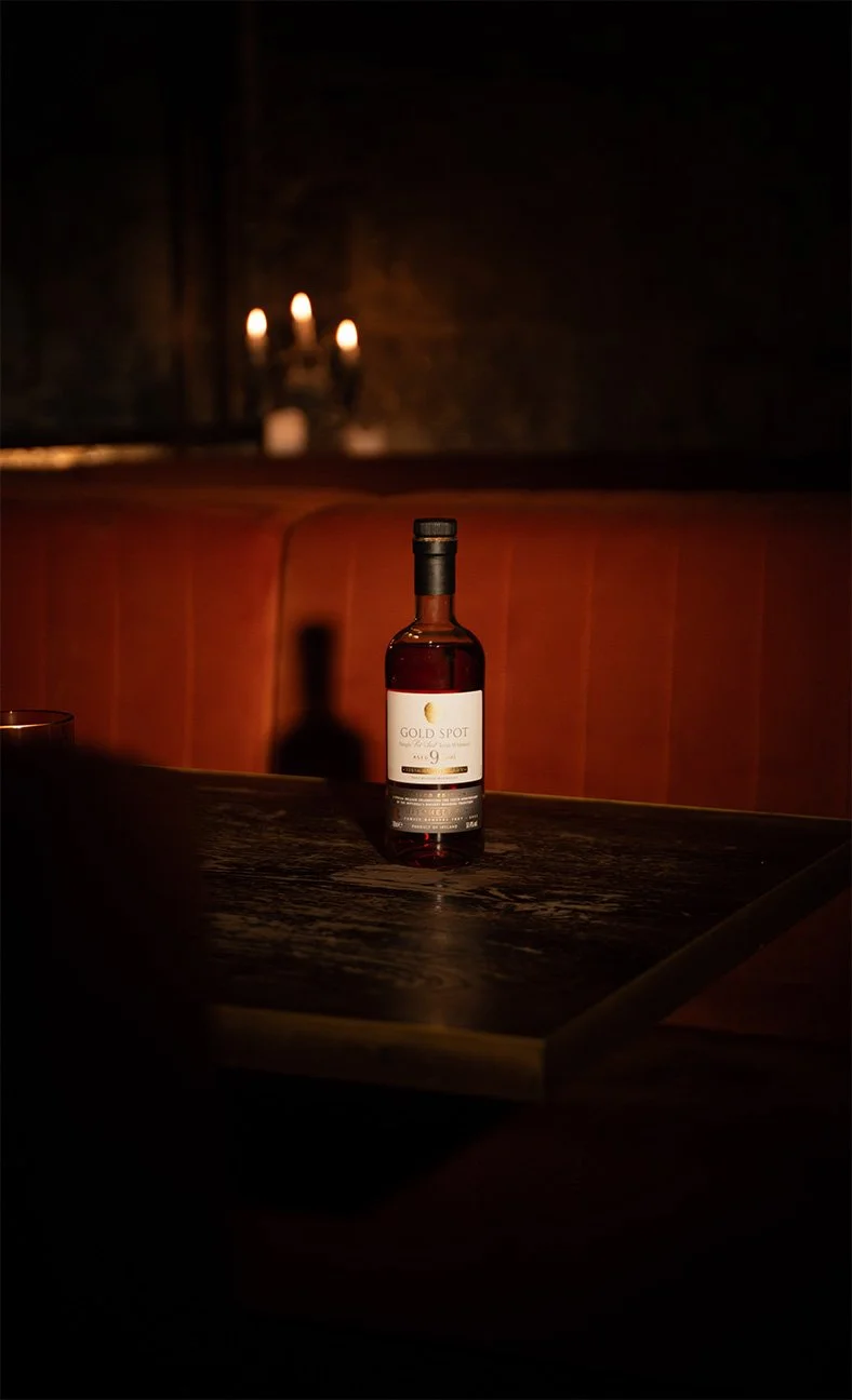 A bottle of Gold Spot Indian whisky on a wooden table in a dimly lit room with a red cushioned bench and a wall sconce with lit candles in the background.