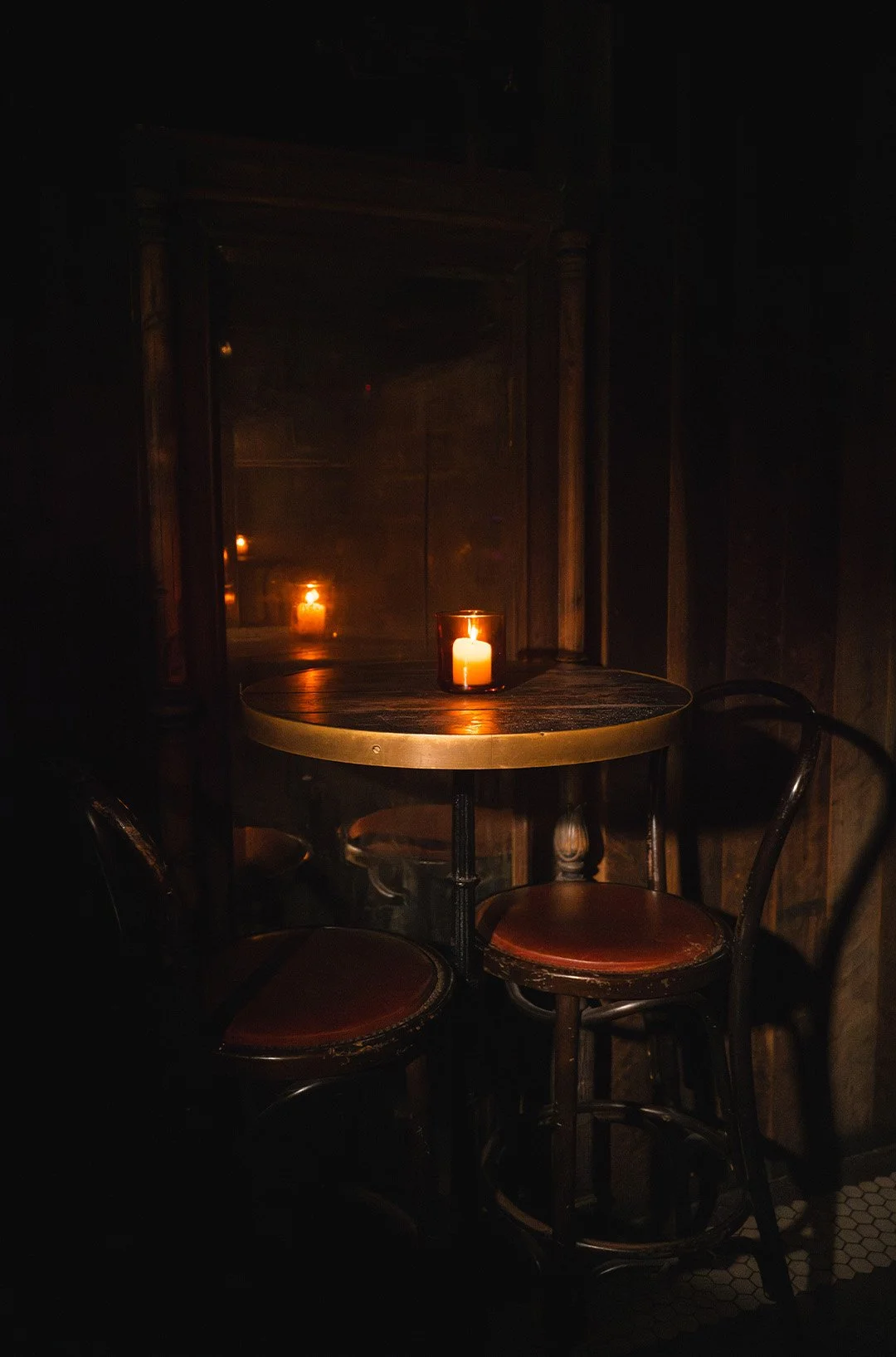 Dimly lit wooden table with a lit candle, reflected in a mirror, surrounded by three wooden chairs in a cozy, dark setting.