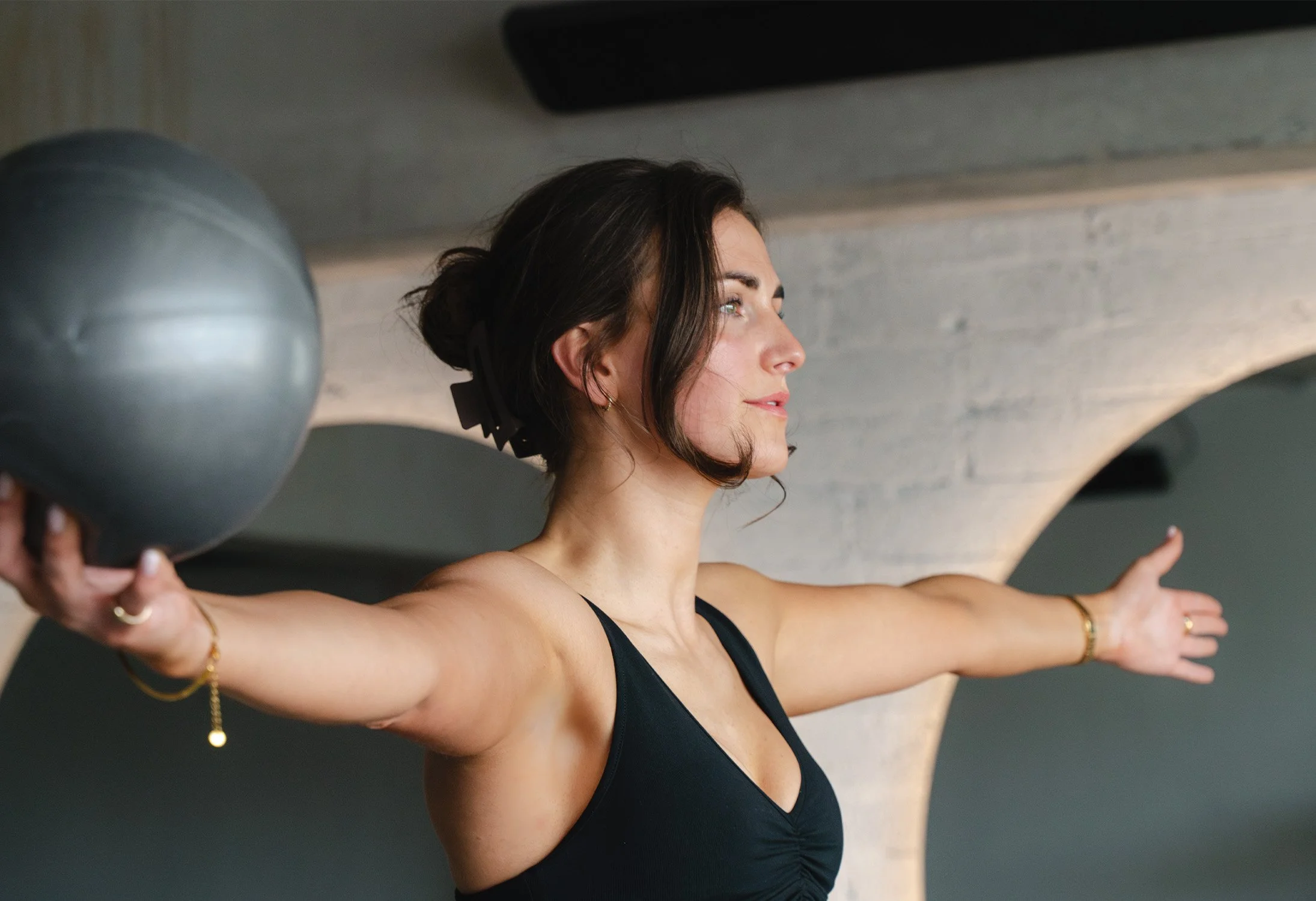 A woman in a black sports bra holding a gray exercise ball with arms outstretched during a workout.