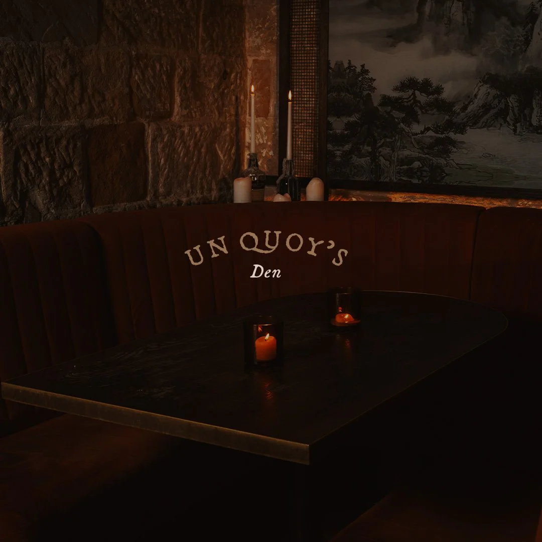 A cozy, dimly lit corner of a restaurant named 'Un Quoy's Den' with candles on the table and candles on the wall. The ambiance features stone and wood decor with artwork in the background.