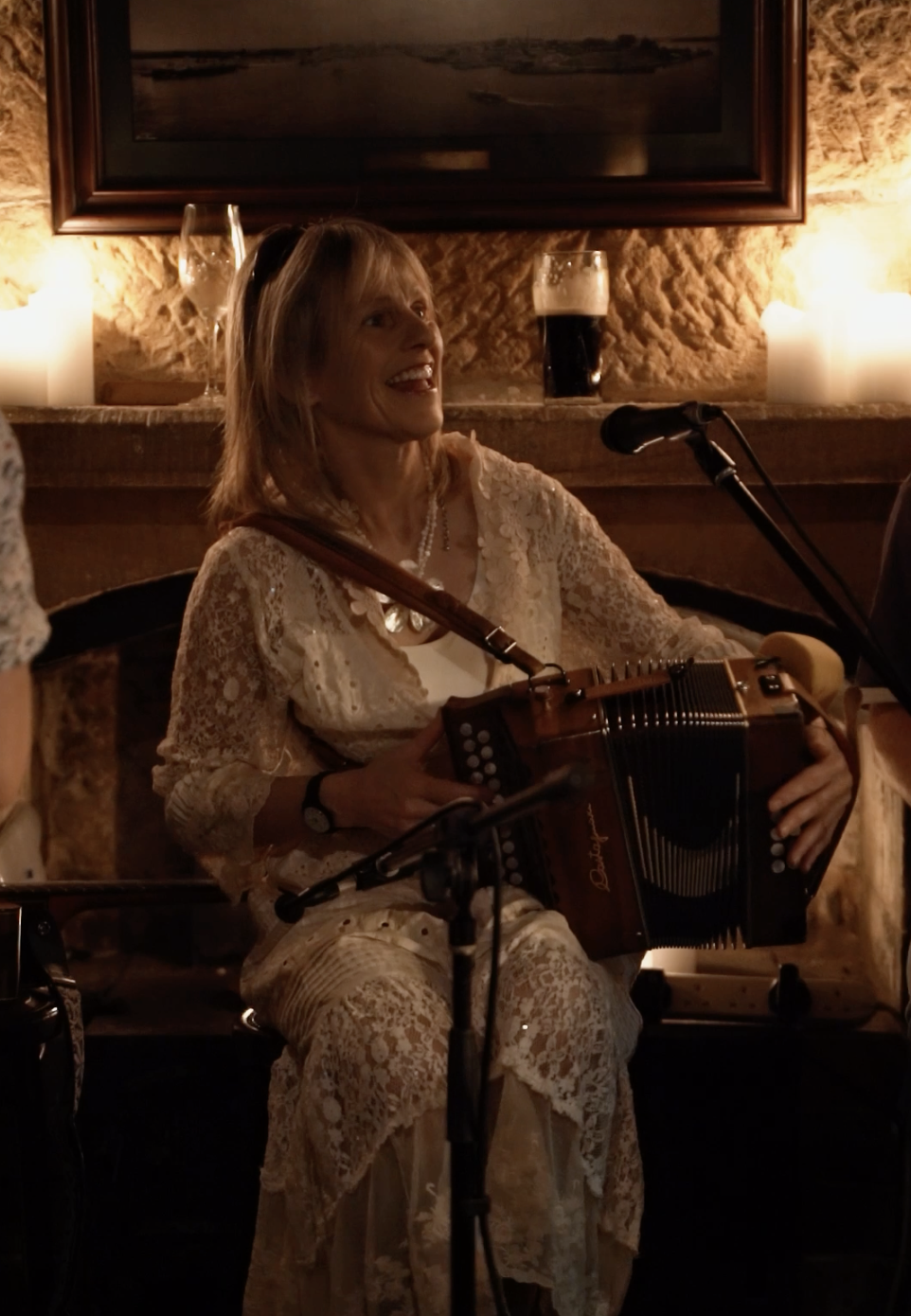 A woman sitting and smiling while playing an accordion during a live performance in a cozy, dimly lit setting with candles, a framed picture, and drinks behind her.