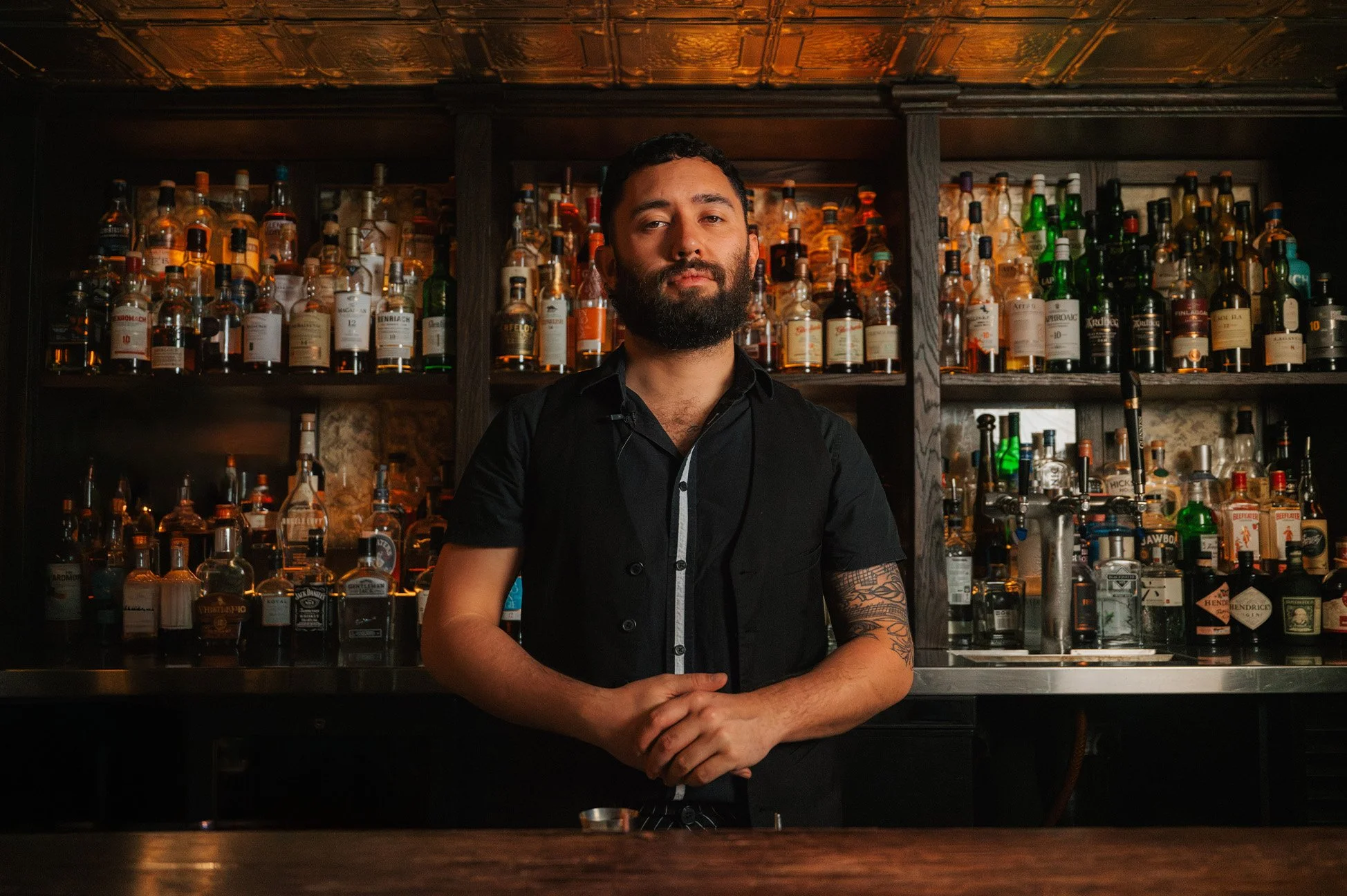 A bearded bartender standing behind a bar with numerous bottles of liquor on shelves in the background.