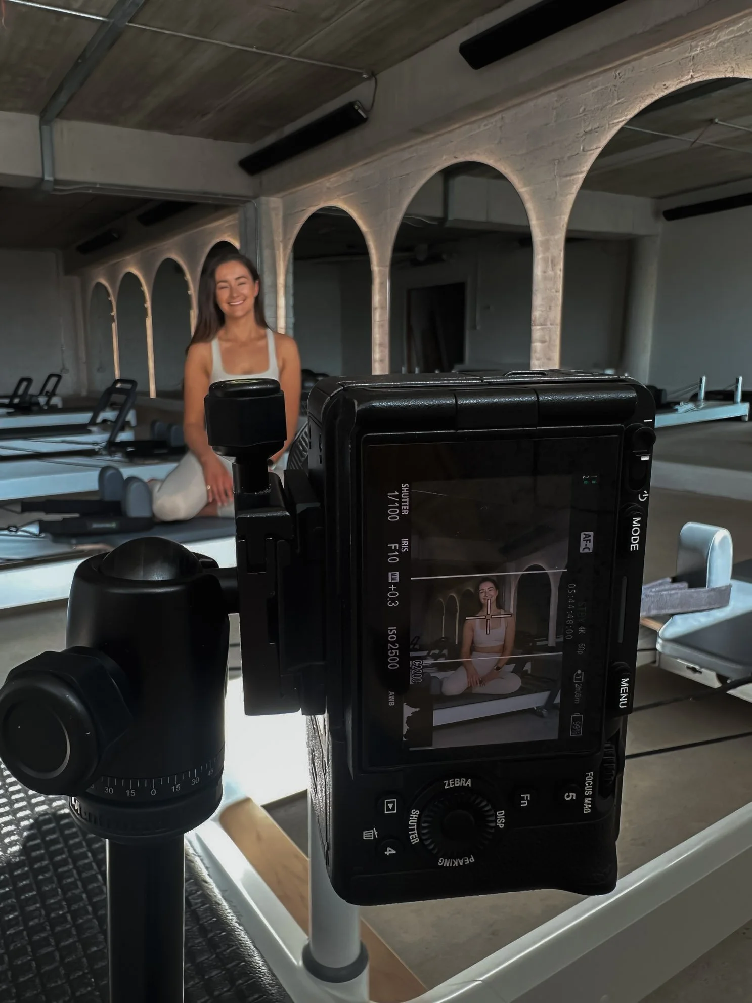 A woman in a white tank top and white pants sitting cross-legged on a Pilates reformer in a fitness studio. She is smiling with her eyes closed, and the photo is being taken with a digital camera mounted on a tripod.
