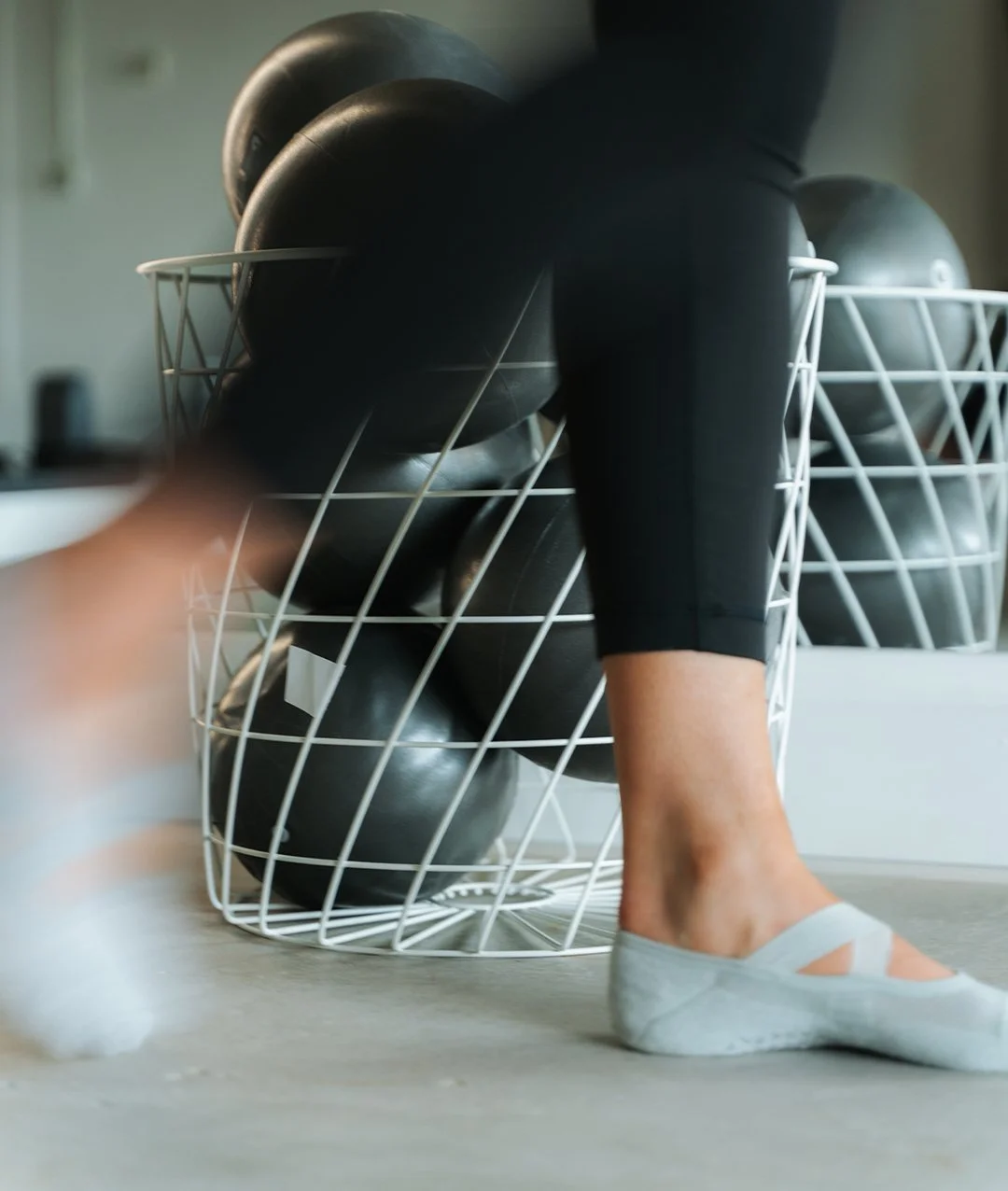 Person wearing gray socks and black workout leggings placing black medicine balls into a white wire basket.