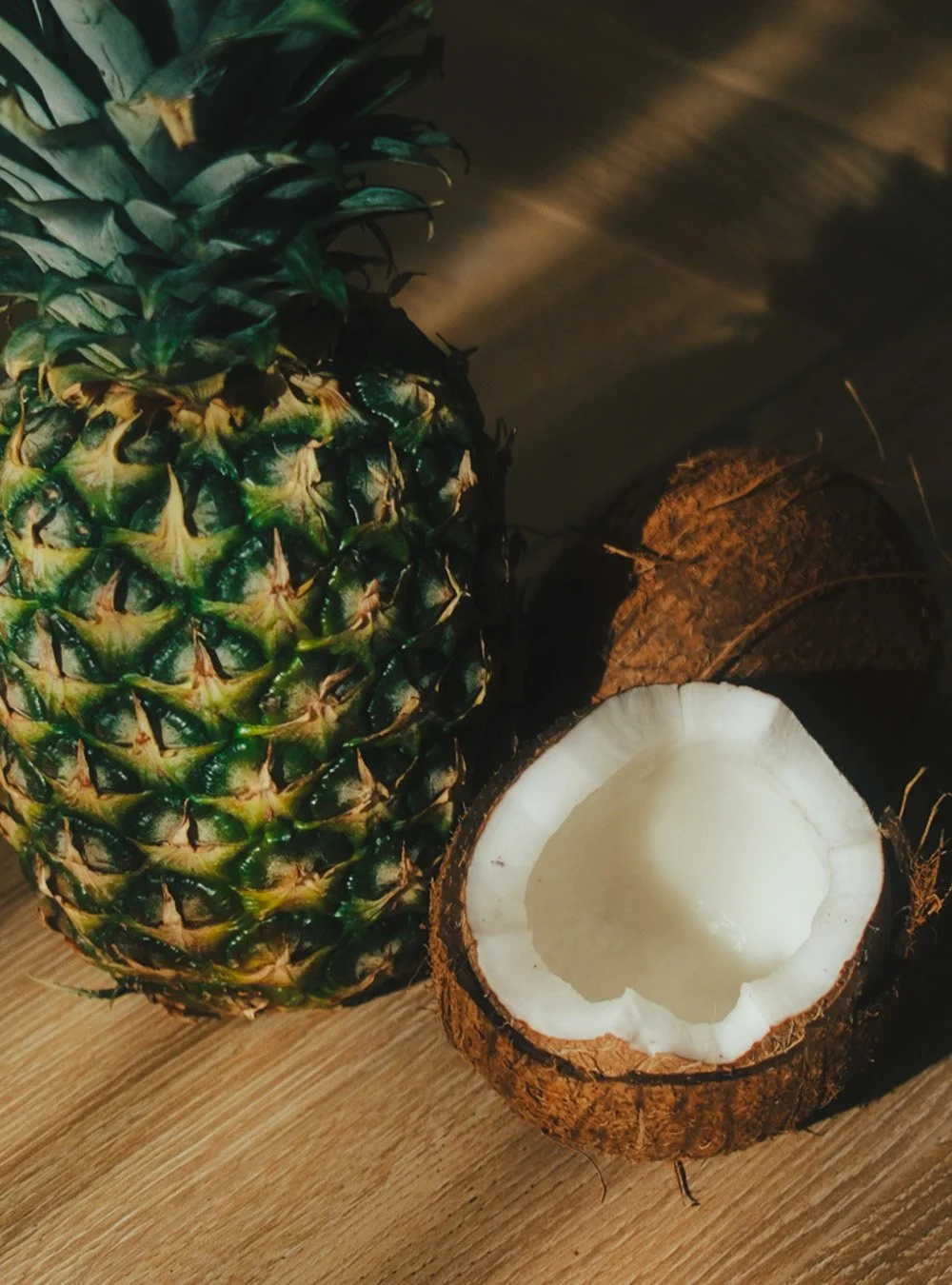 A whole pineapple next to a halved coconut with white flesh, on a wooden surface.