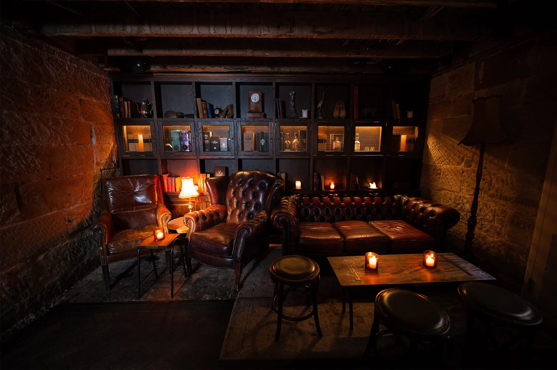 Cozy dimly lit lounge with leather armchairs and a sofa, candles on tables, dark wooden bookshelf with decorative items and books, exposed brick and stone walls, ambient lighting.