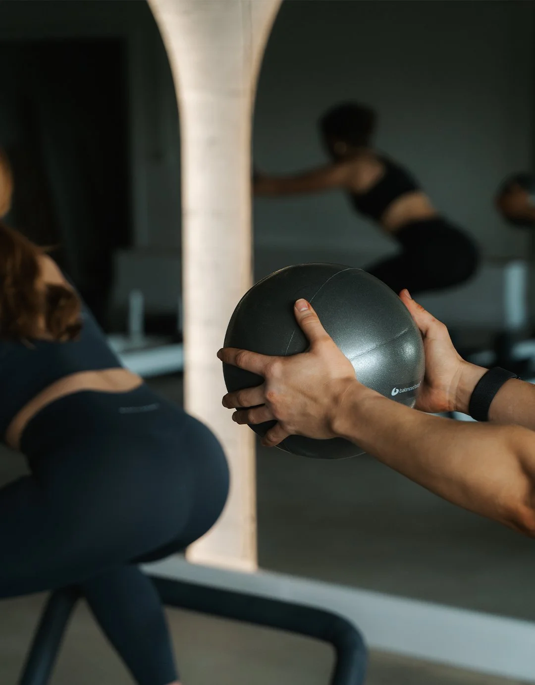 Person holding a black exercise ball during a workout in a gym, with a mirror reflecting a woman performing squats in the background.