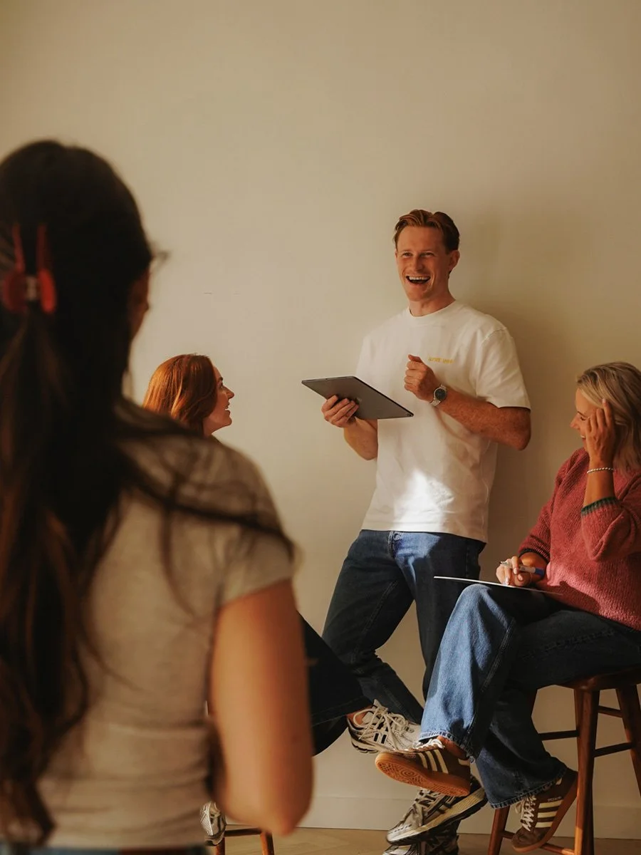 A group of four people having a discussion, with one man standing and speaking, holding a tablet, while three women are seated and listening, in a casual setting.