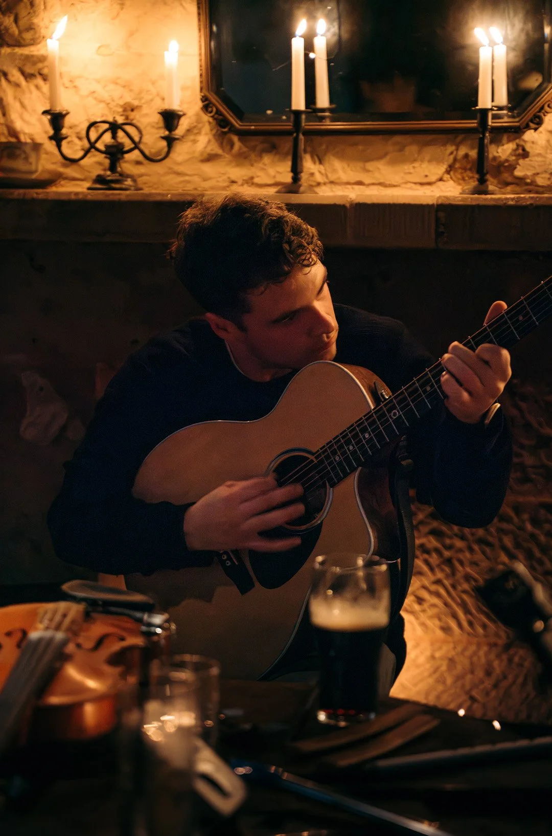 Young man playing an acoustic guitar in a cozy, dimly-lit room with a stone wall, candles on the mantle, and a mirror above.