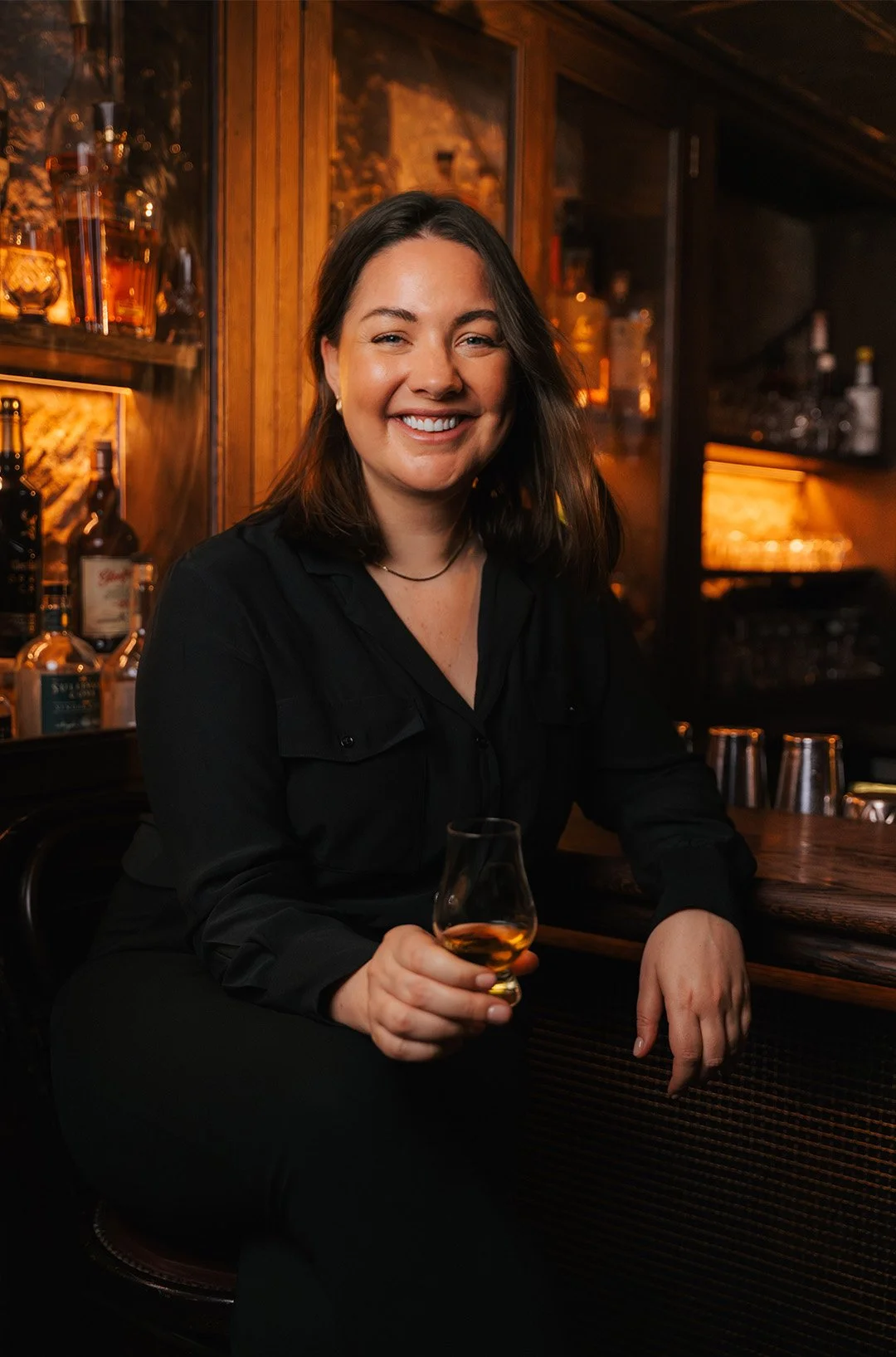 A woman smiling and sitting at a bar, holding a glass of whiskey.
