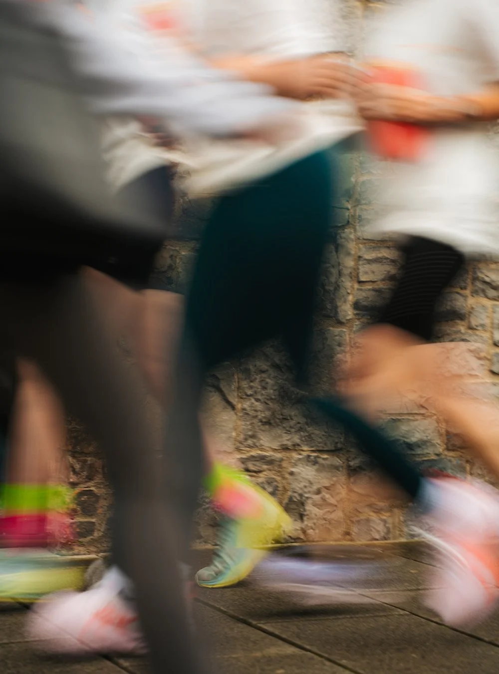 Blurred motion image of people running in athletic shoes near a brick wall, with visible legs and feet.
