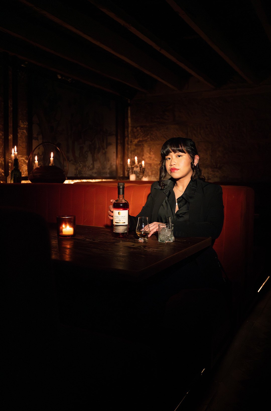 A woman in a black outfit sitting at a wooden table in a dimly lit room, holding a bottle of whiskey, with several glasses and candles on the table, a candlestick and more candles in the background.