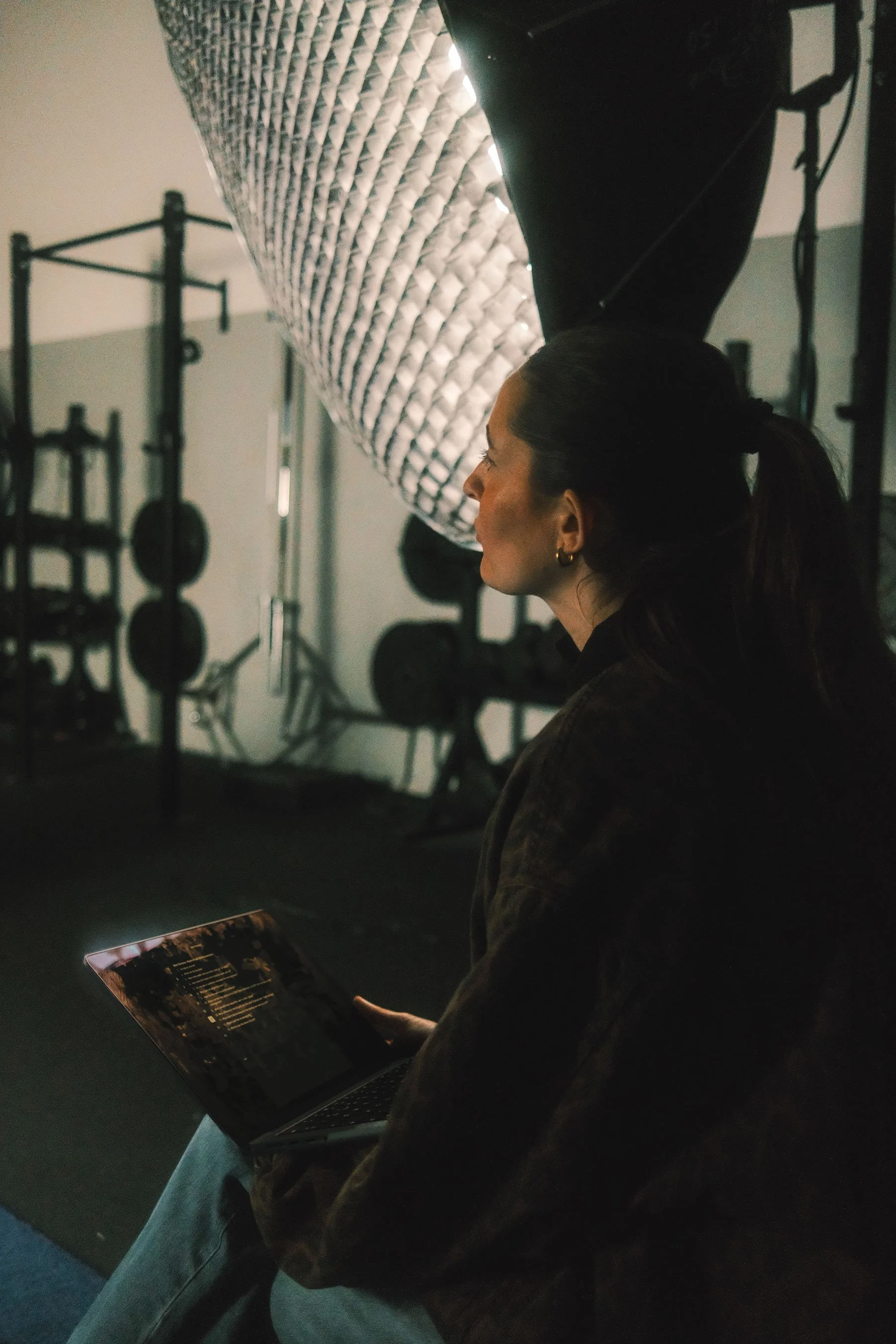 A woman sitting in a gym, holding a tablet, with studio lighting equipment and gym equipment in the background.