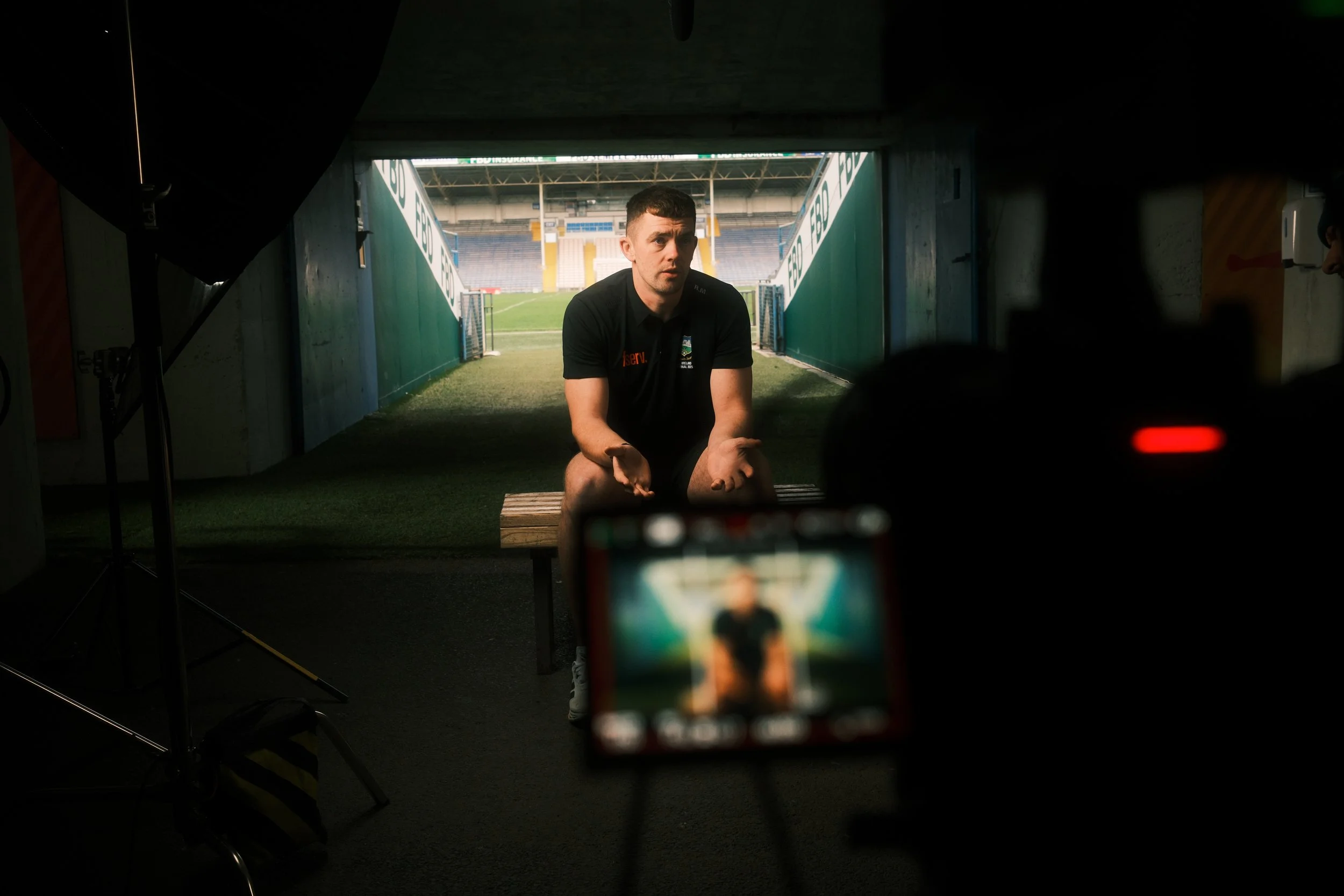 A man sitting on a bench inside a tunnel leading to a sports field, being filmed or photographed with a camera and lighting equipment in a dimly lit room.
