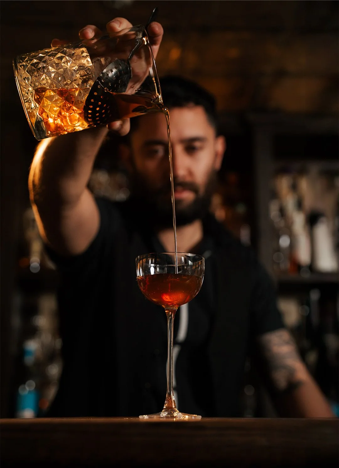 Bartender pouring dark liquor into a cocktail glass at a bar, with bar shelves in the background.