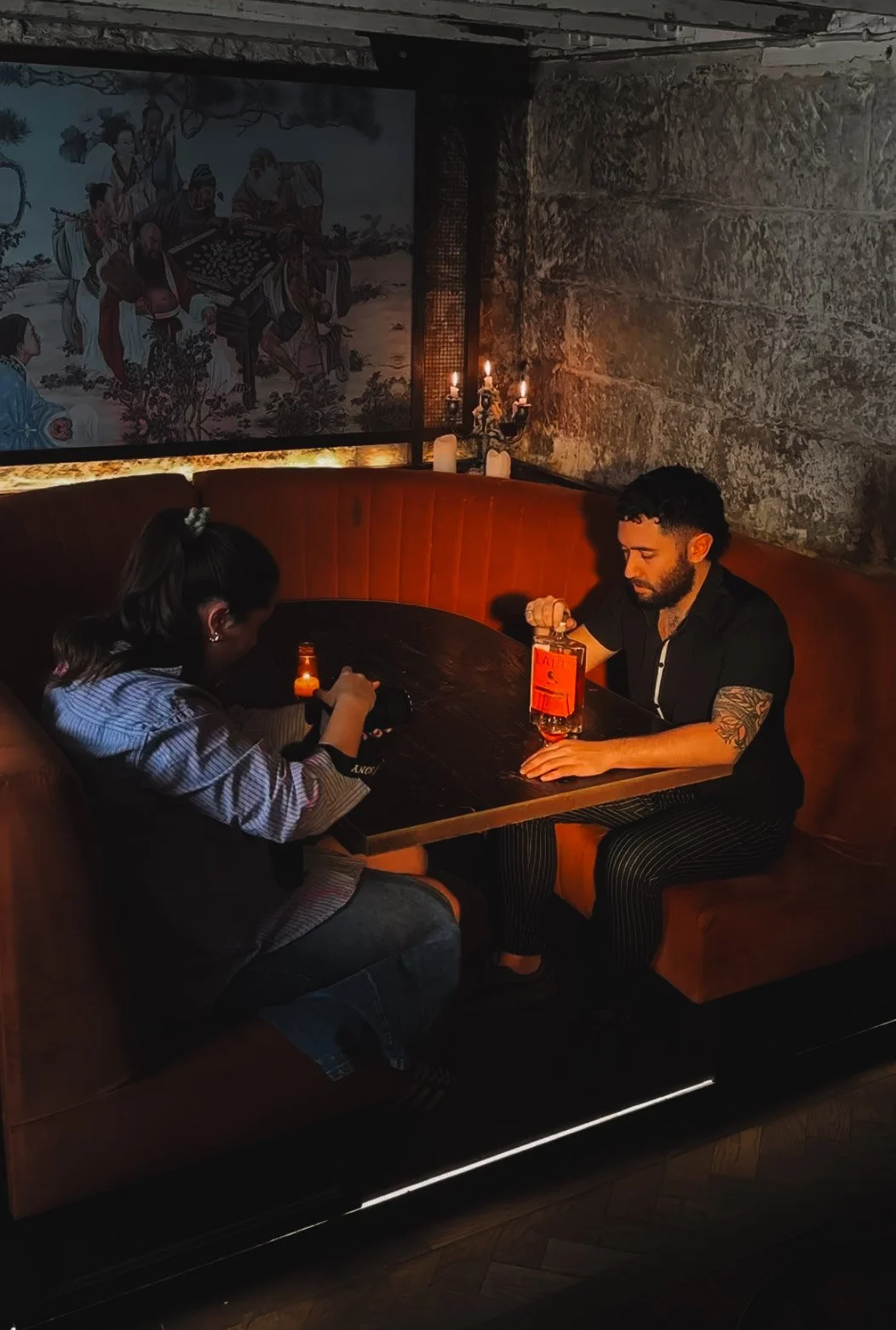 A young man with tattoos on his arm sitting at a restaurant table with a young girl, taking a photo of a bottle of alcohol, with a dimly lit background, stone walls, a painting on the wall, and candles.