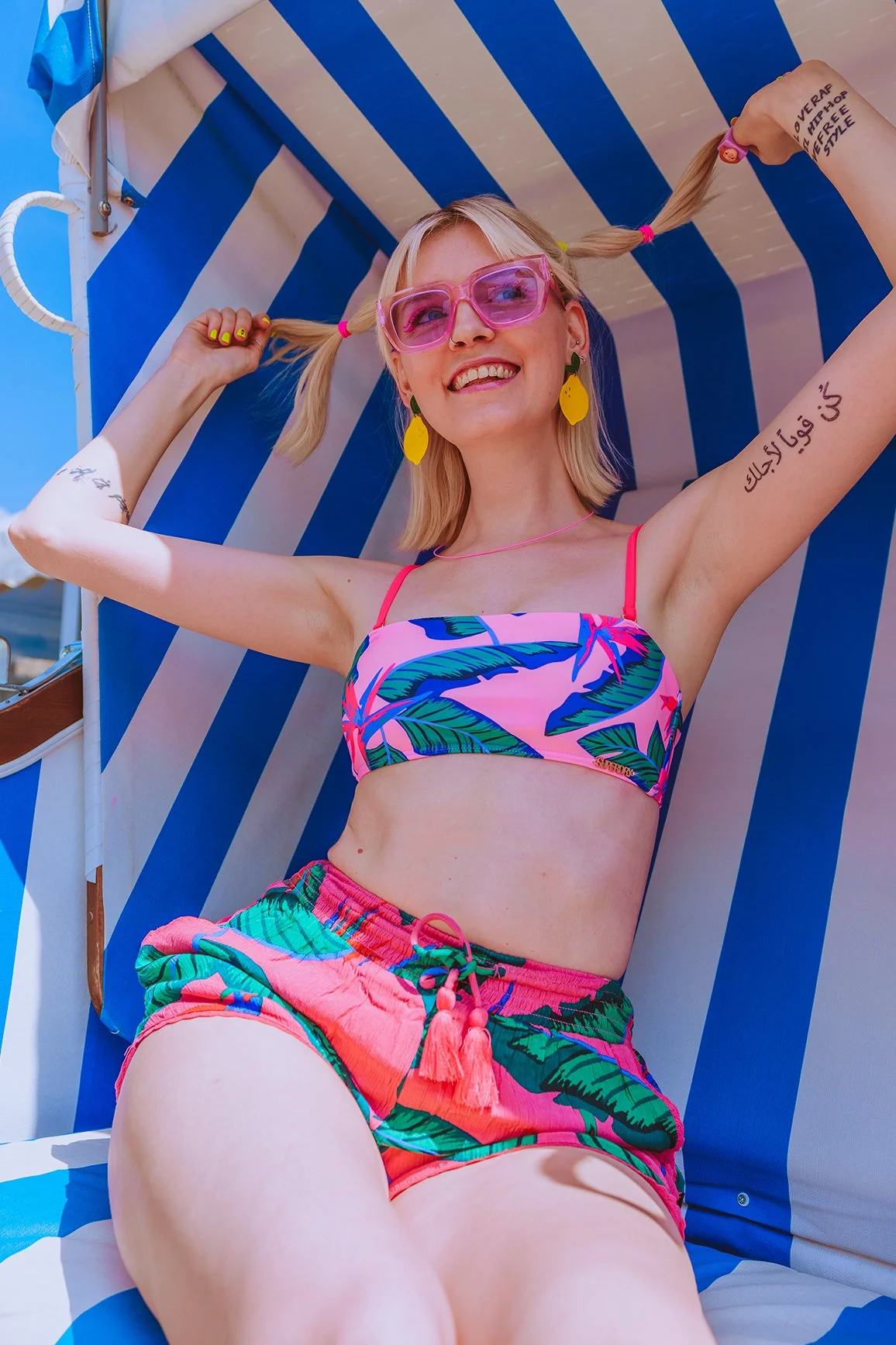 A woman with blonde hair and pink sunglasses is sitting on a blue and white striped outdoor lounge chair. She is smiling and playfully holding her braided hair with both hands. She is wearing a colorful pink and green tropical-patterned bikini top and matching shorts, with yellow leaf-shaped earrings. The background shows a sunny day at the beach or poolside.