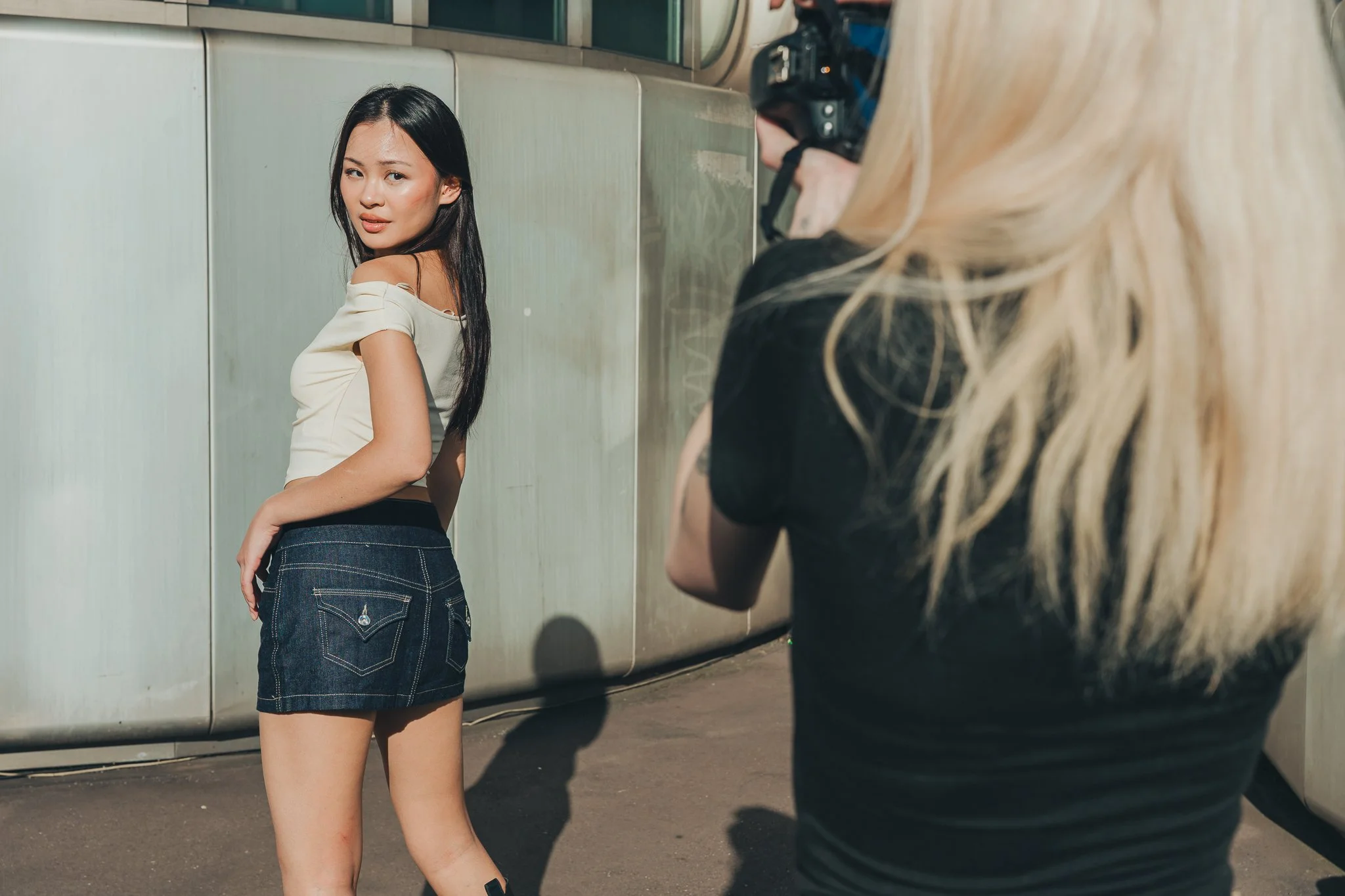 A woman with long black hair wearing a beige off-shoulder top and denim skirt posing outdoors while a photographer captures her picture.