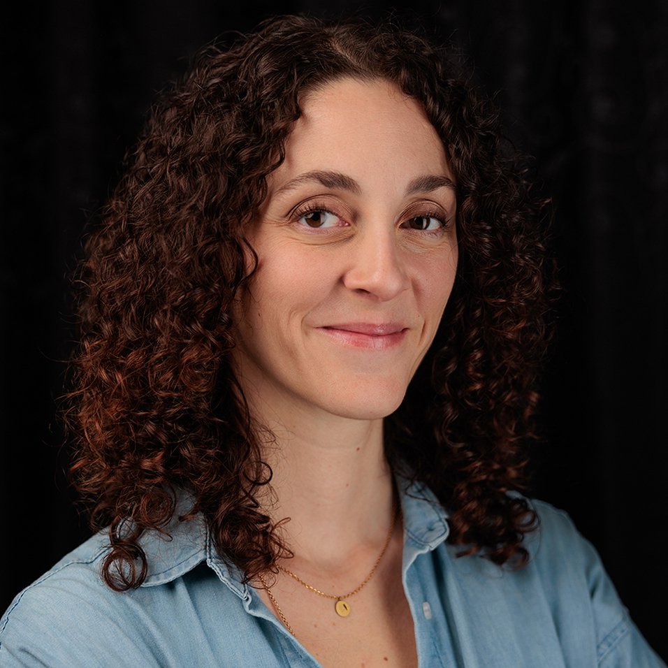 A woman with curly brown hair smiling at the camera against a dark background.
