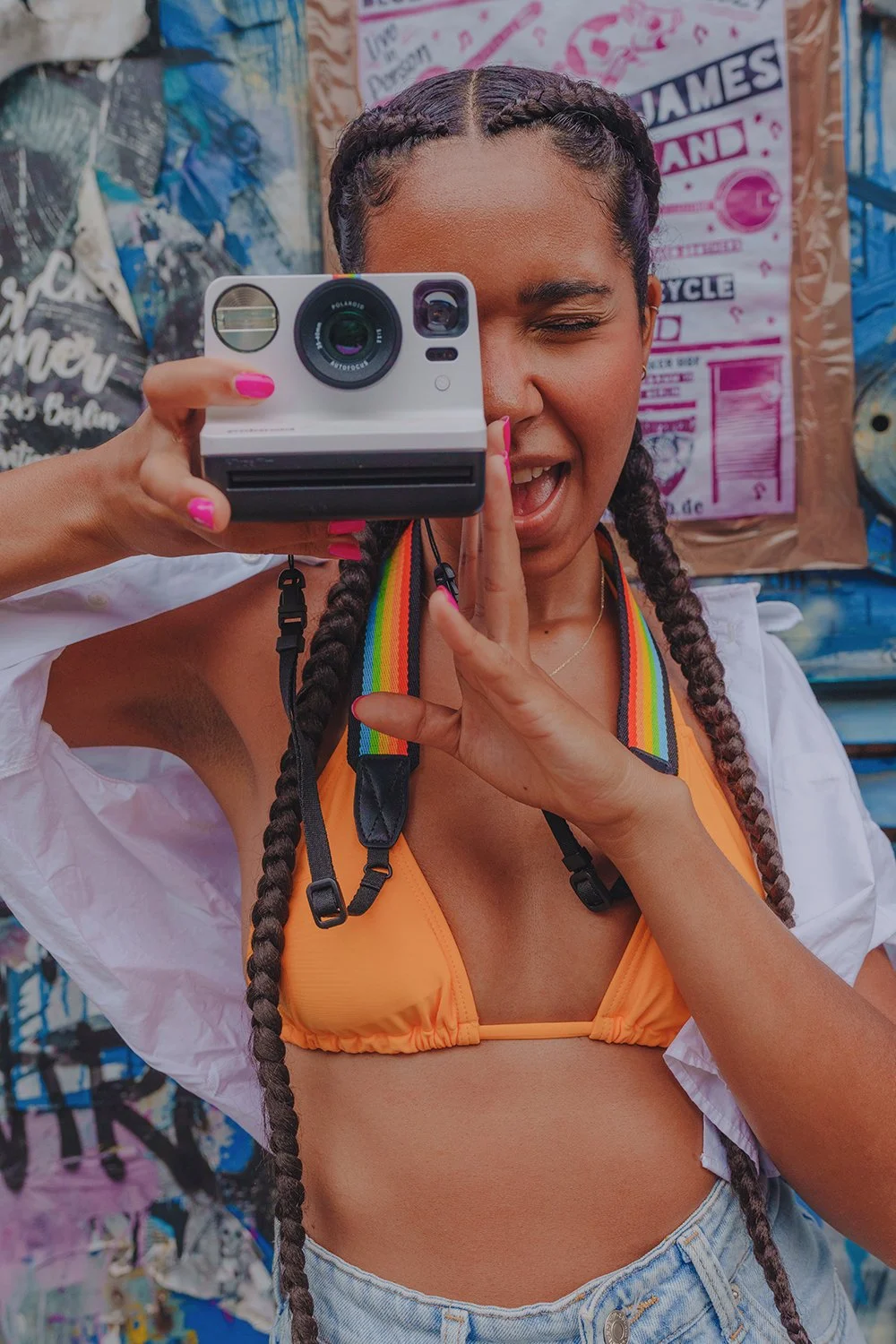 Woman in a bikini top with braided hair, taking a photo with a Polaroid camera, smiling and winking, against a colorful graffiti background.