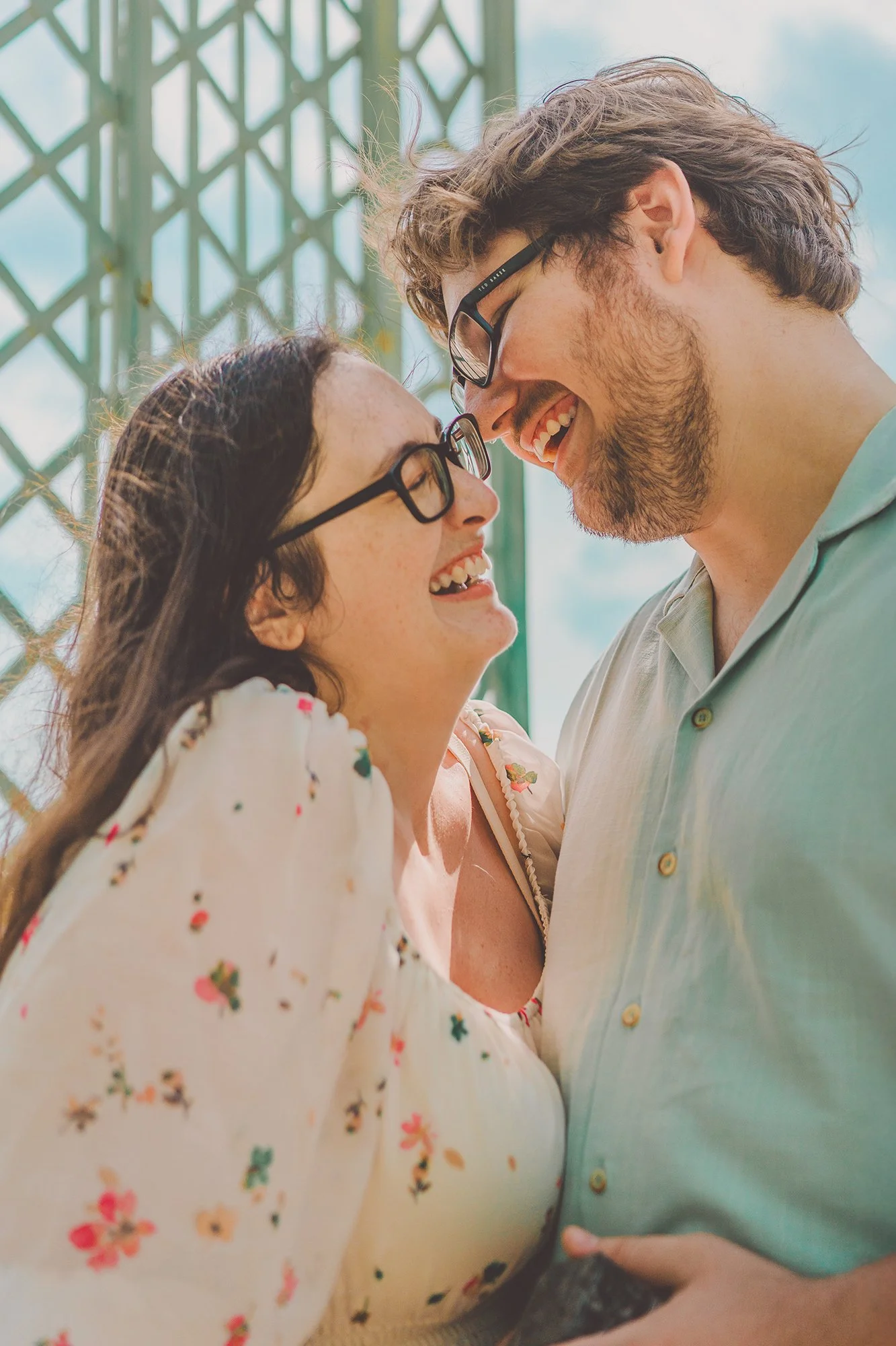 A smiling young woman and a smiling young man with glasses are close together, almost touching foreheads, outdoors near a green lattice structure.