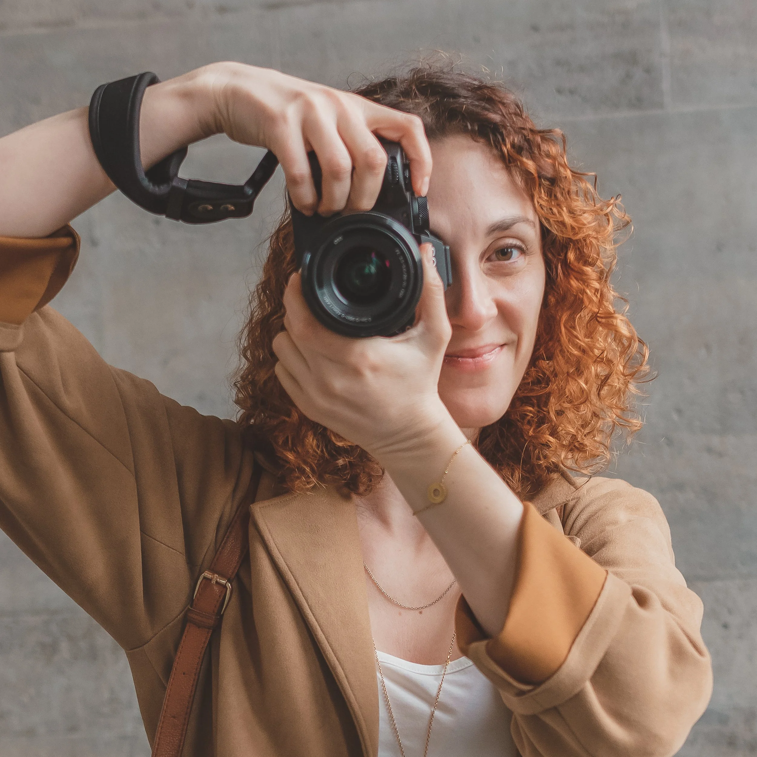 A woman with curly red hair taking a photo with a camera indoors against a gray wall.