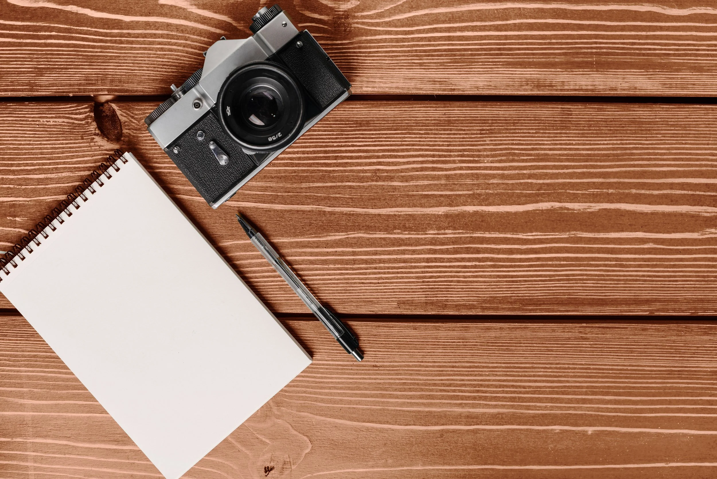 A flat lay of a vintage camera, a black and clear pen, a blank spiral notebook, and a brown wooden surface.