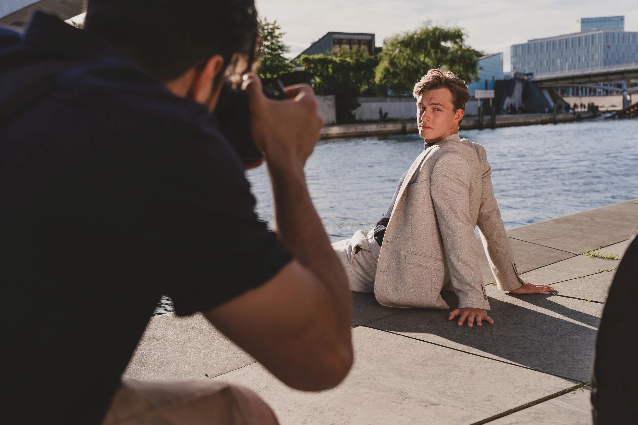 A young man in a beige blazer sitting on a concrete ledge by a river, looking at the camera, while a photographer kneels in front taking his picture.