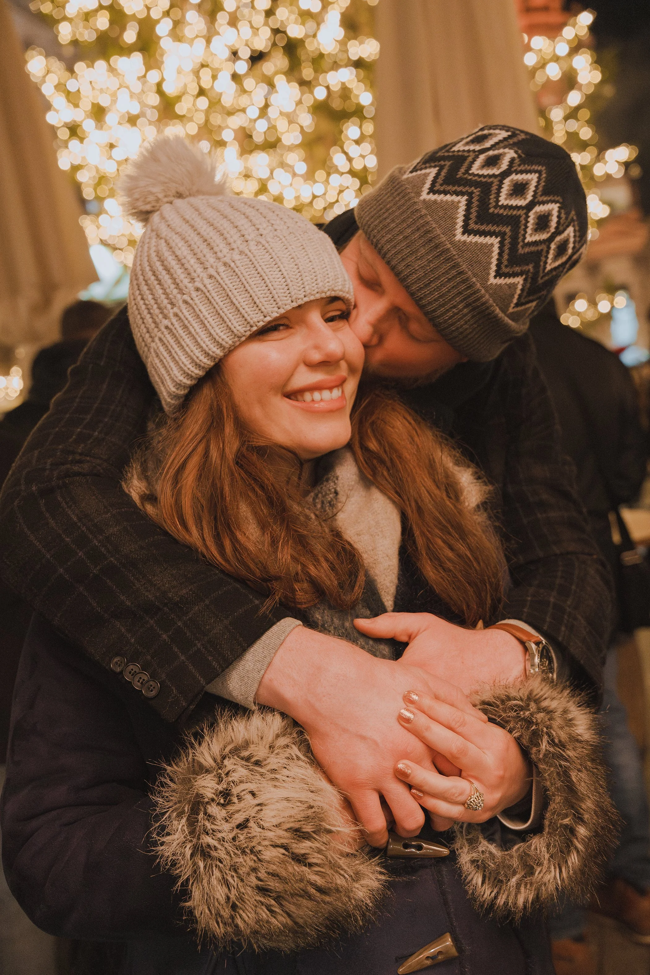 A couple dressed warmly in winter clothing exchanging a kiss and hug in front of a decorated Christmas tree with lights.