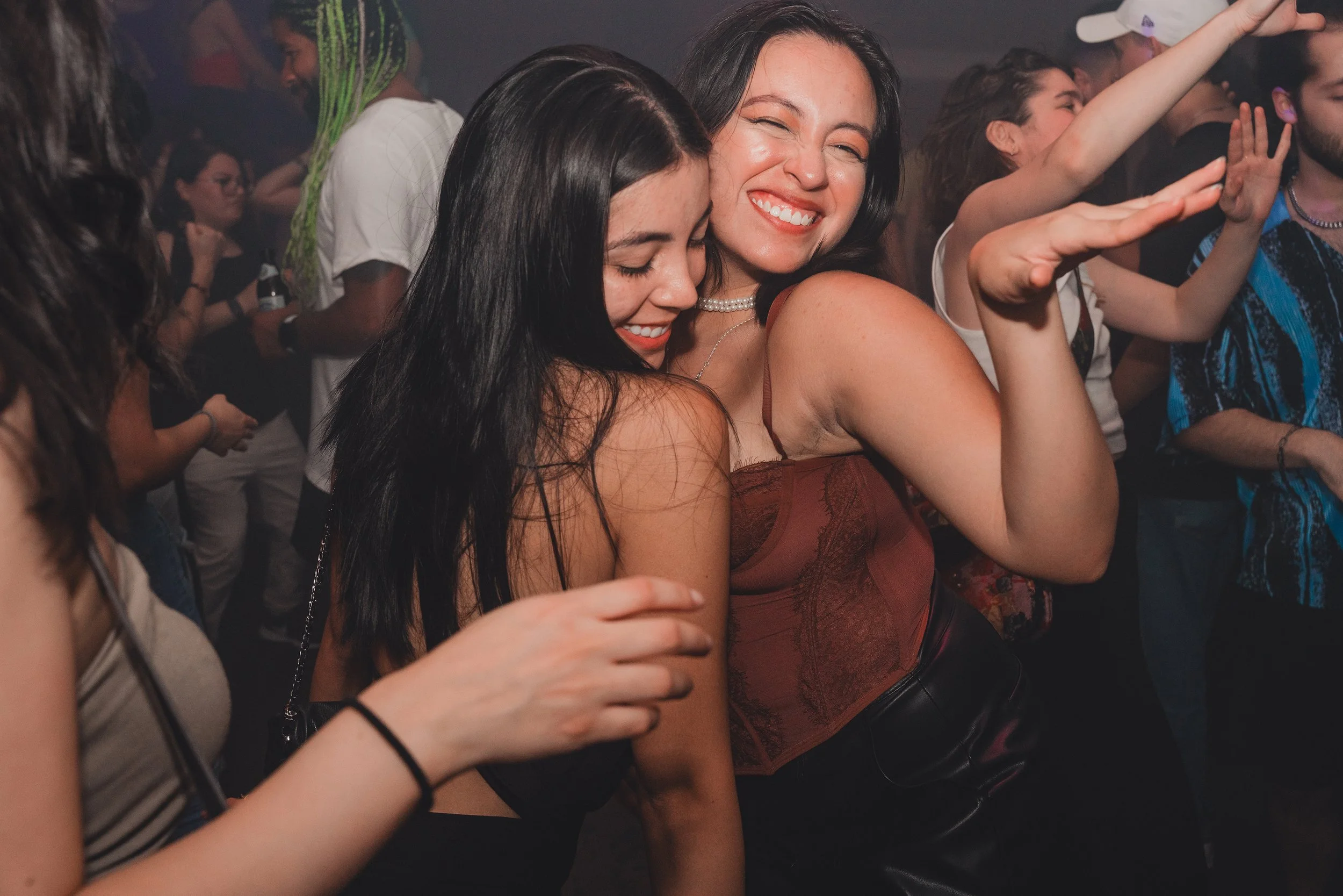 Two women are dancing and smiling at a party or nightclub, surrounded by other people, with their arms raised and enjoying the moment.