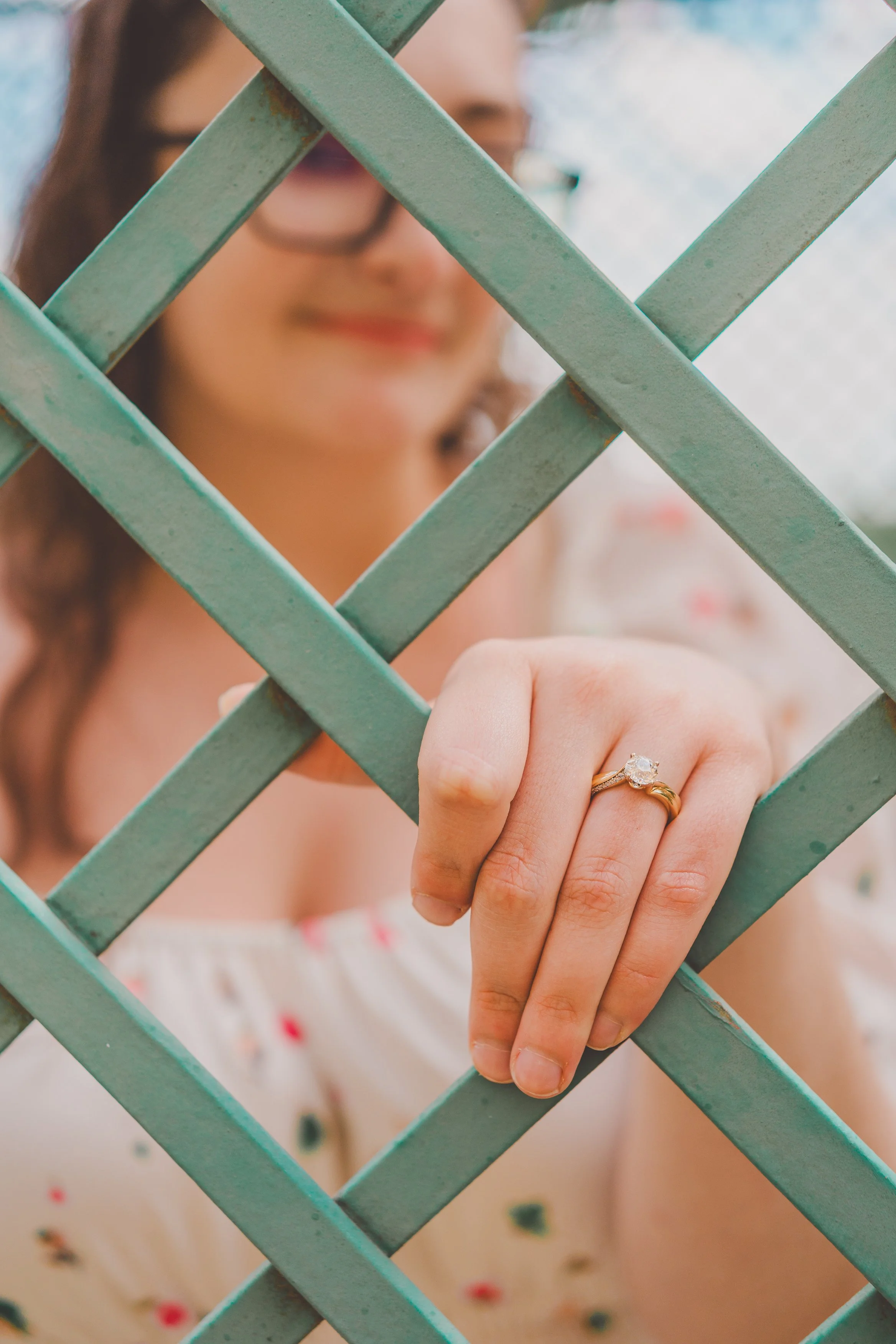 A woman wearing a ring on her finger, holding onto a teal lattice fence. The woman is smiling and dressed in a light-colored dress with small floral prints.