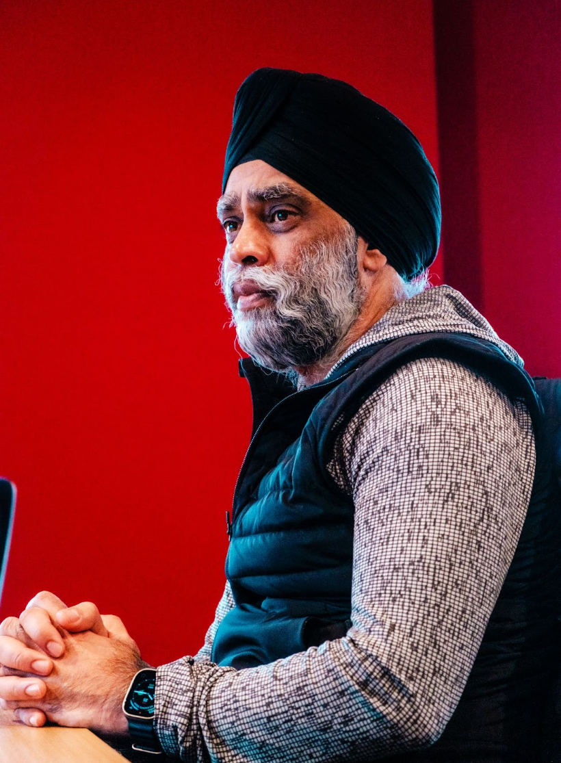 A middle-aged man wearing a black turban, a checkered shirt, and a black vest looks attentively to the side against a red background.
