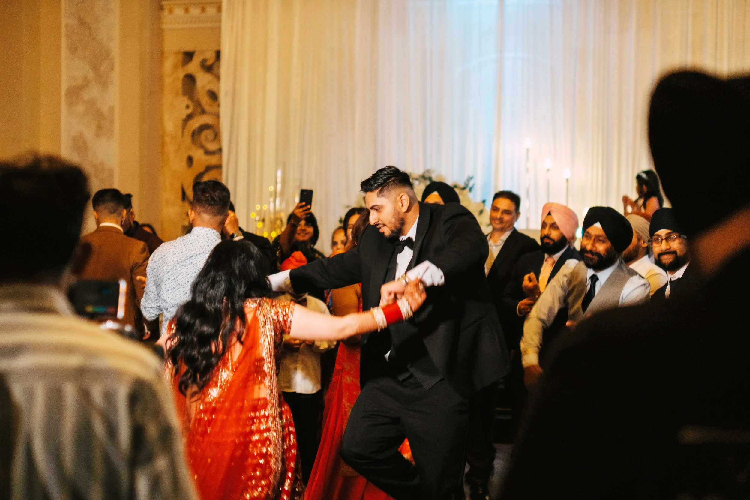 A man in a tuxedo dancing with a woman in a red Indian dress at a wedding reception. They are holding hands and dancing on a decorated dance floor while surrounded by guests, some of whom are taking photos and watching.