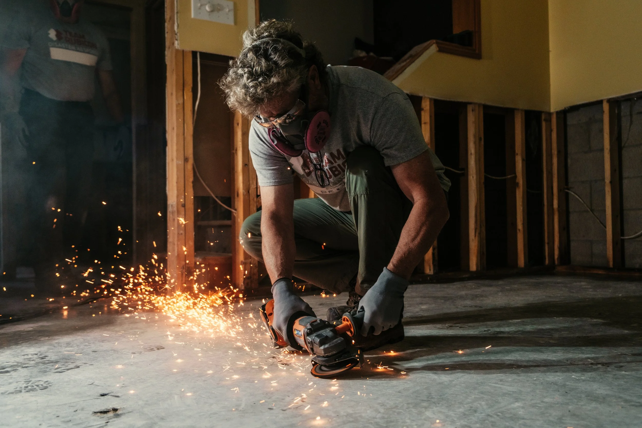 A person wearing safety gloves, a t-shirt, and a face mask is crouched on the floor operating a power tool, with sparks flying around, in an unfinished room with wooden framing and exposed electrical wiring.