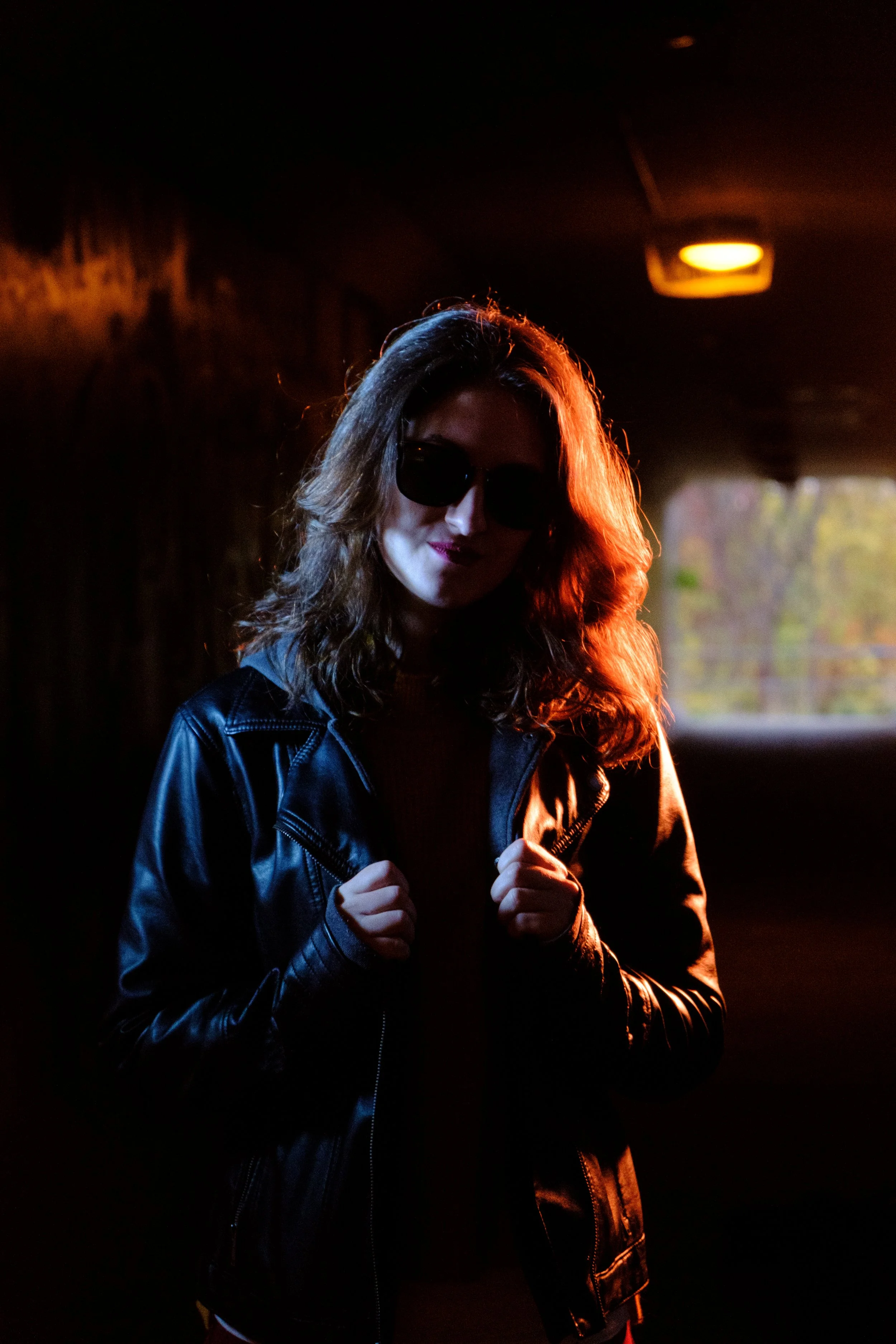 A woman with wavy hair wearing sunglasses and a leather jacket, standing in a dark parking garage with orange lighting.