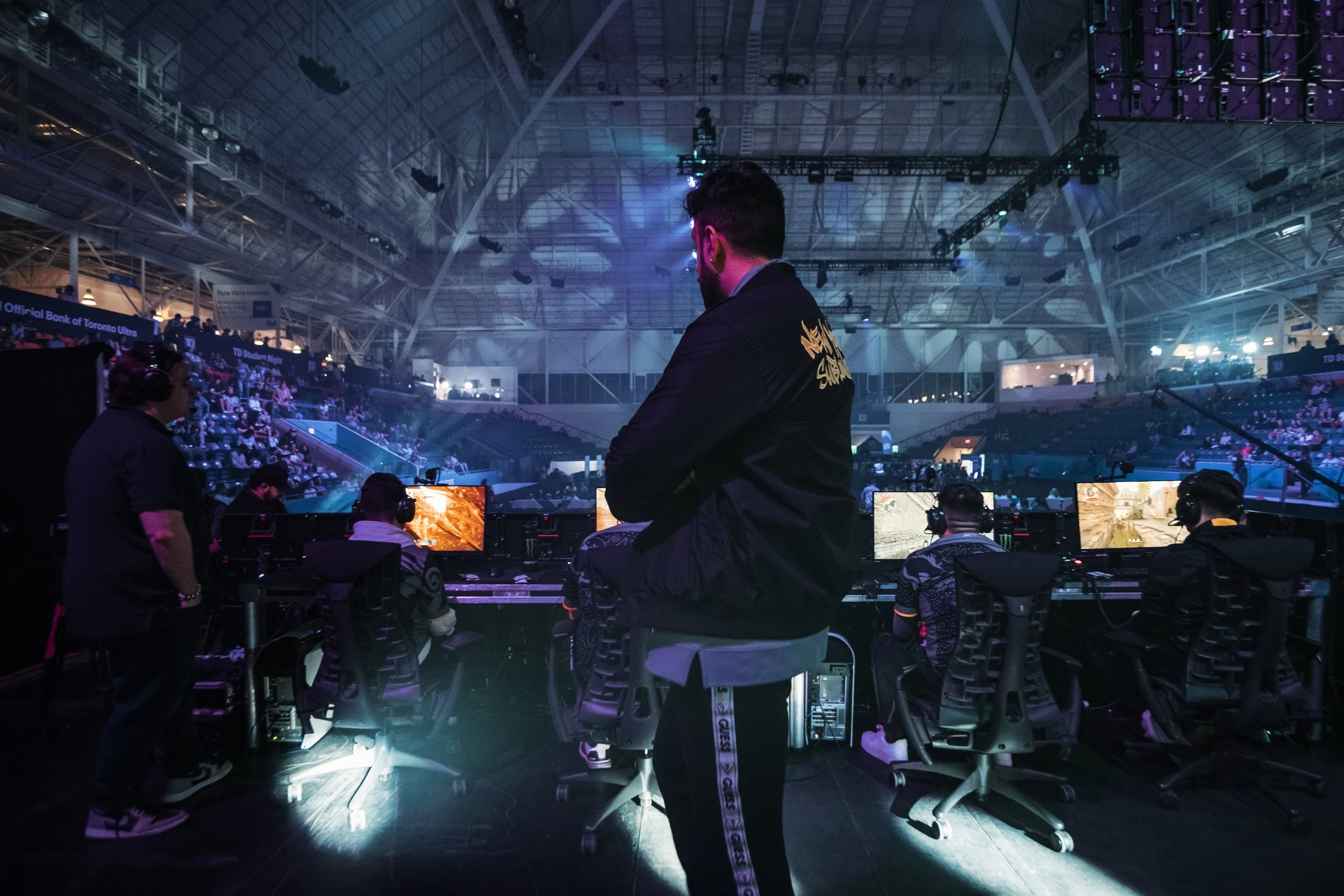 An esports event with players seated at gaming stations on a stage, and with an audience watching from the stands in a large, illuminated indoor arena.