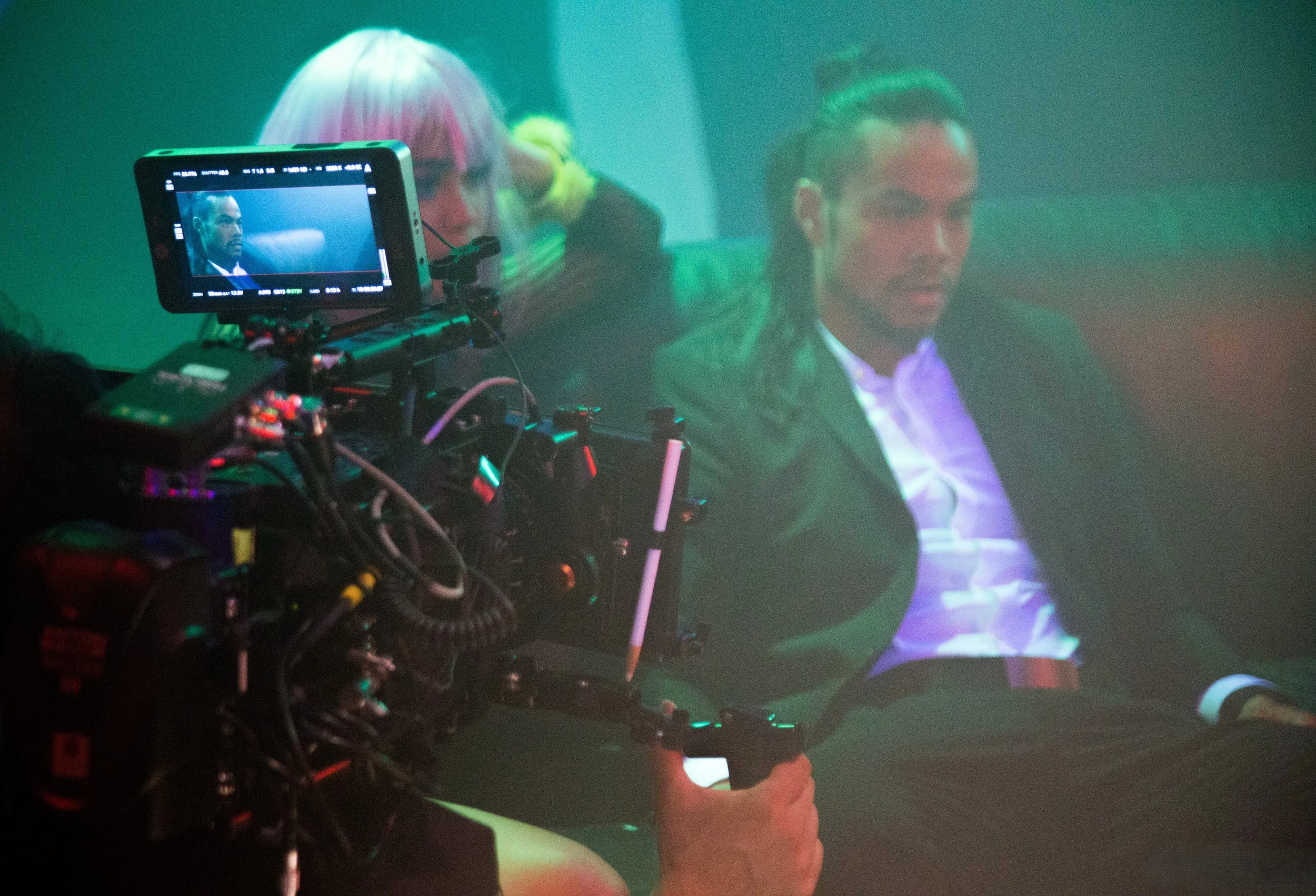 A film set scene with a camera focused on a man with long hair, sitting in a chair, wearing a dark blazer and white shirt. The camera is on a dolly and shows the scene being recorded on its screen.