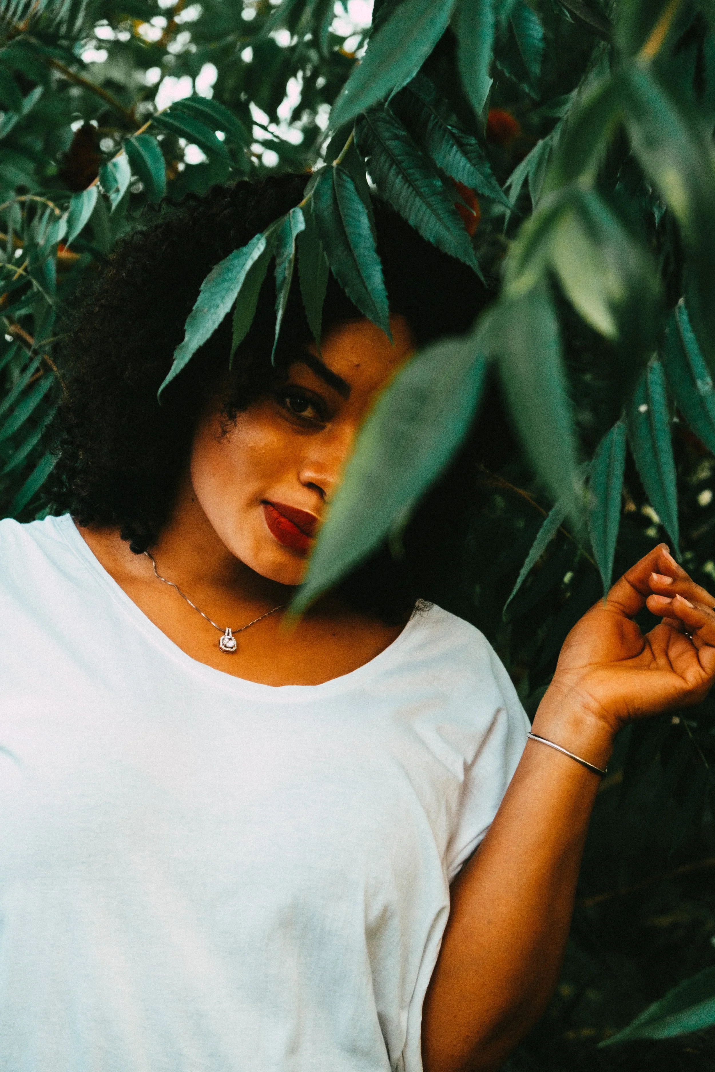 A woman with curly hair wearing a white shirt, jewelery, and red lipstick among green leaves. She is partially hidden behind the foliage, with a confident expression.