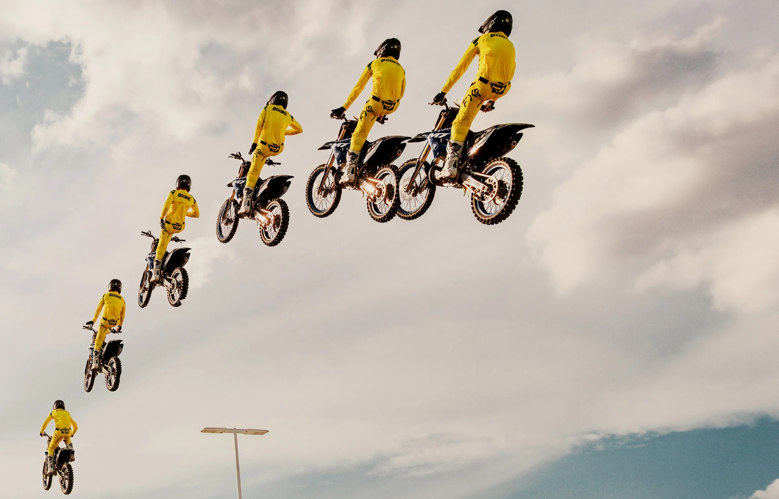 Sequence of six motorcyclists in yellow riding gear performing jumps against a cloudy sky.