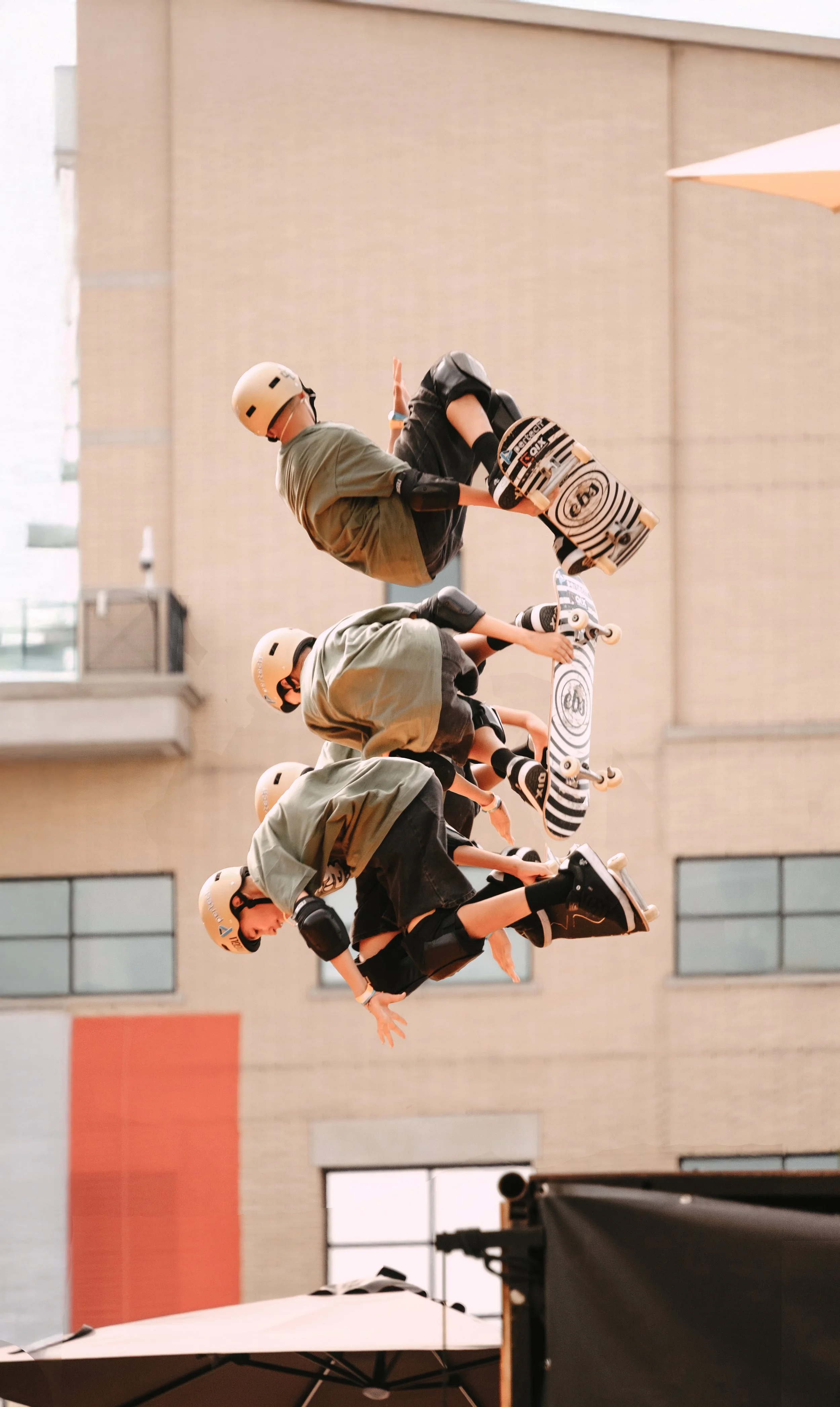 Four skateboarders mid-air performing tricks, all wearing helmets and protective gear, against a backdrop of an urban building.