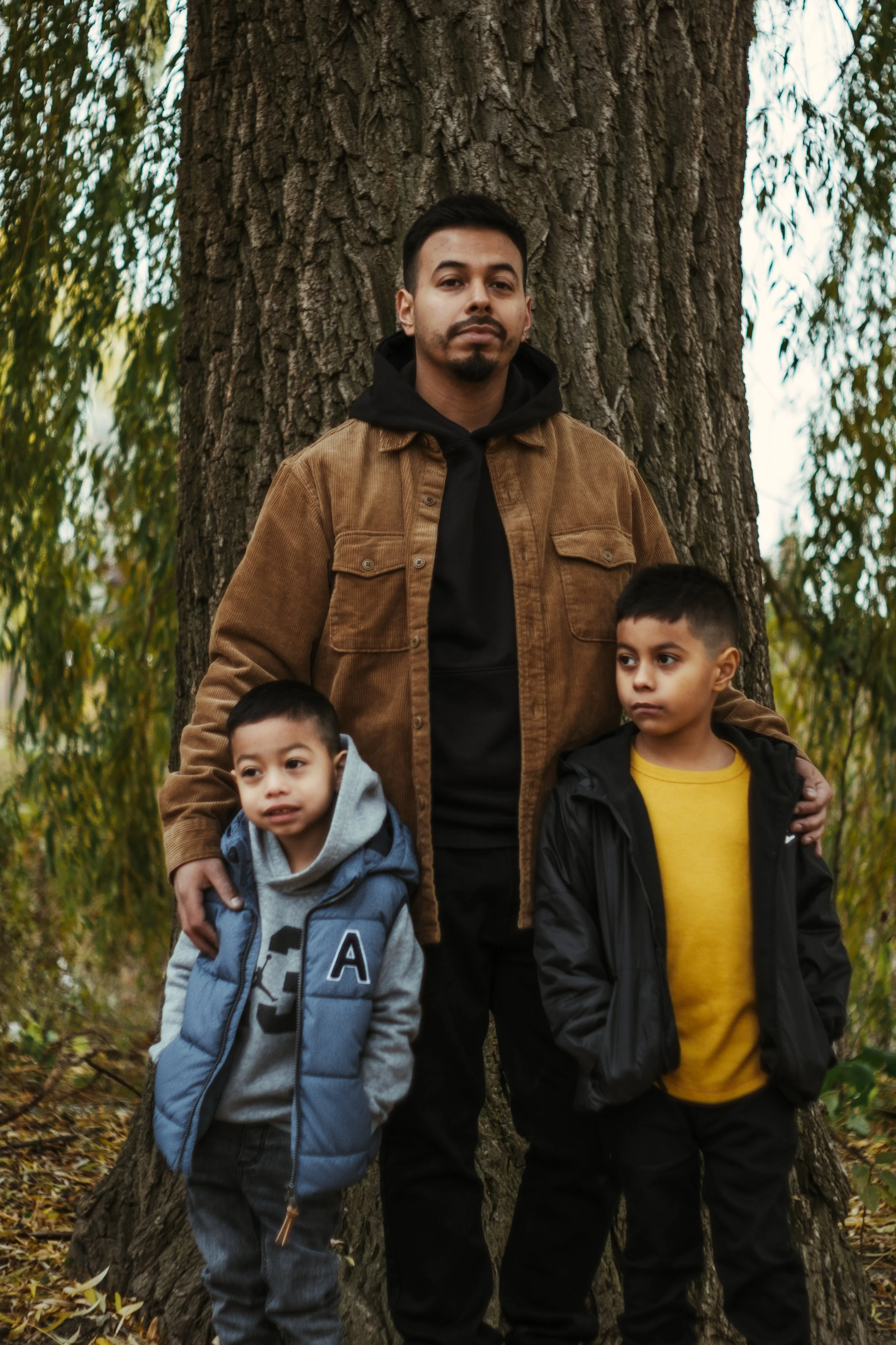 A man standing behind two young boys outdoors in front of a large tree, with greenery and fallen leaves around them.