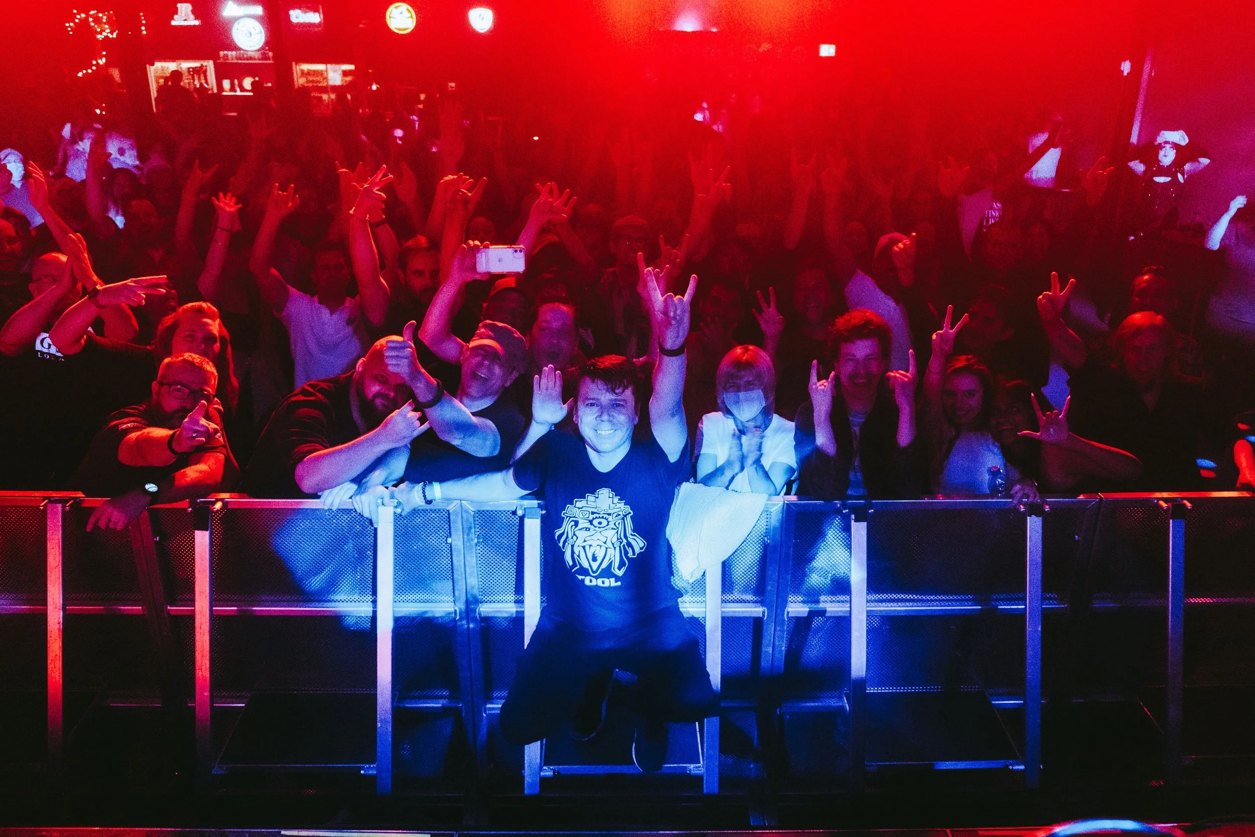 Crowd of concert-goers at a music event, front row smiling, some flashing peace signs, with stage lights casting red and blue hues, and a person in the center wearing a black T-shirt with a lion face graphic.